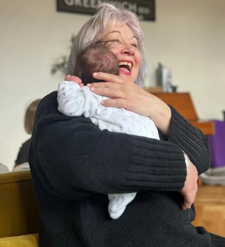 An elderly woman with gray hair smiling and hugging a newborn baby in a cozy indoor setting.