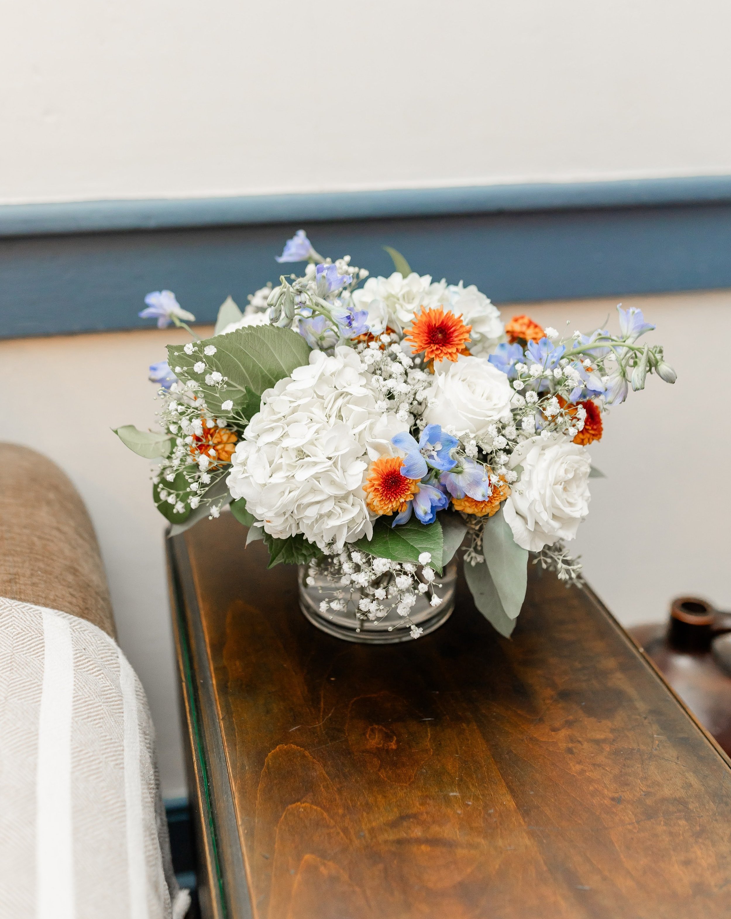 Floral arrangement with white roses, hydrangeas, orange and blue flowers in glass vase on wooden table.