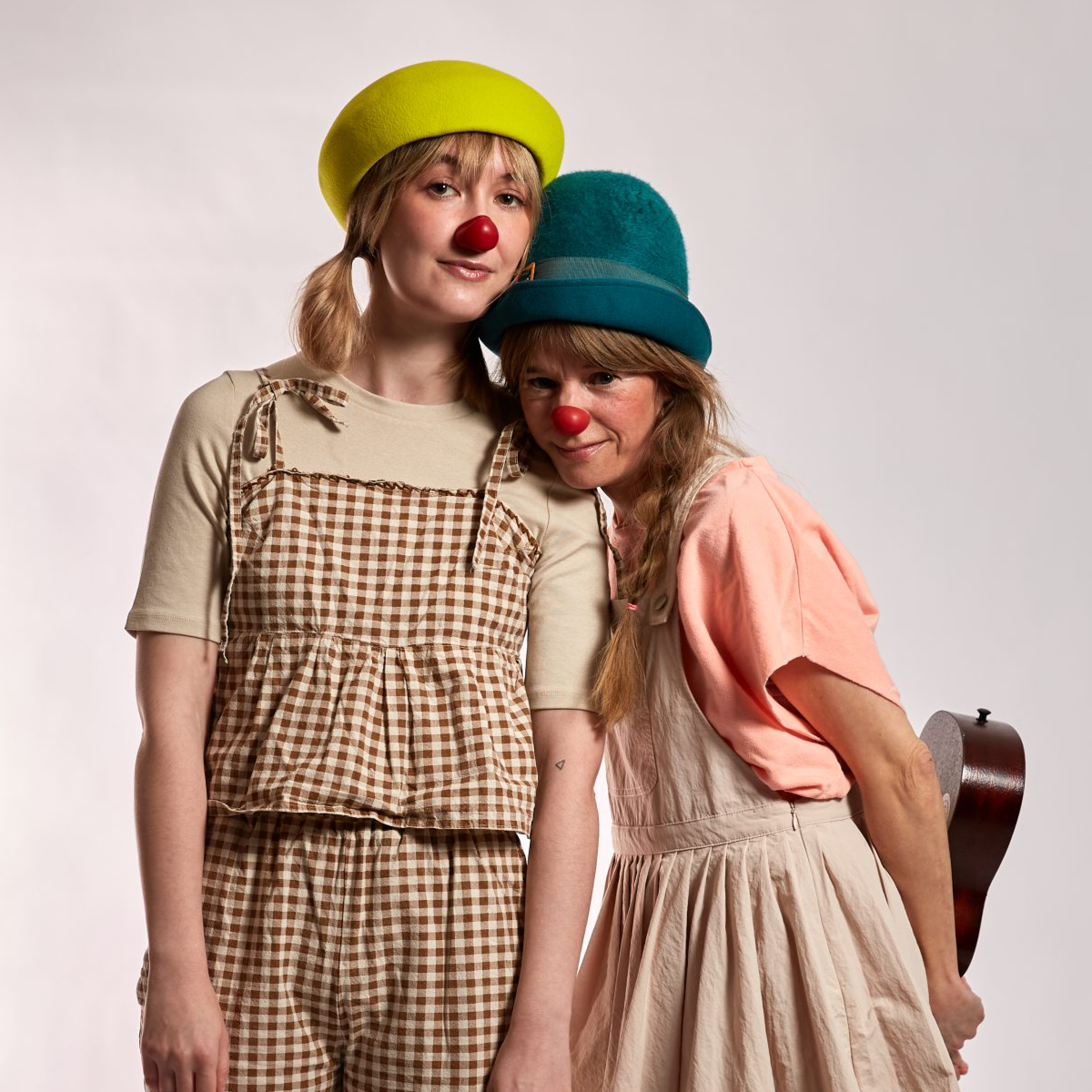 Two women dressed as clowns, wearing colorful hats and red noses, standing close together against a plain light background. One woman has a green hat and a checkered apron, while the other has a blue hat and is holding a guitar.