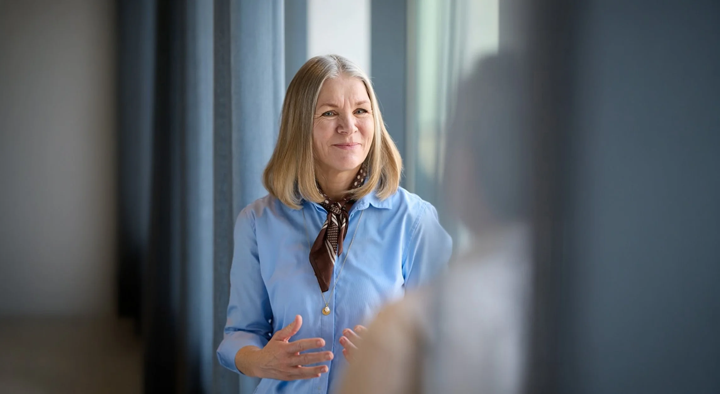 A middle-aged woman with blonde hair, wearing a blue shirt and a brown scarf, smiling and talking in a well-lit room with curtains.