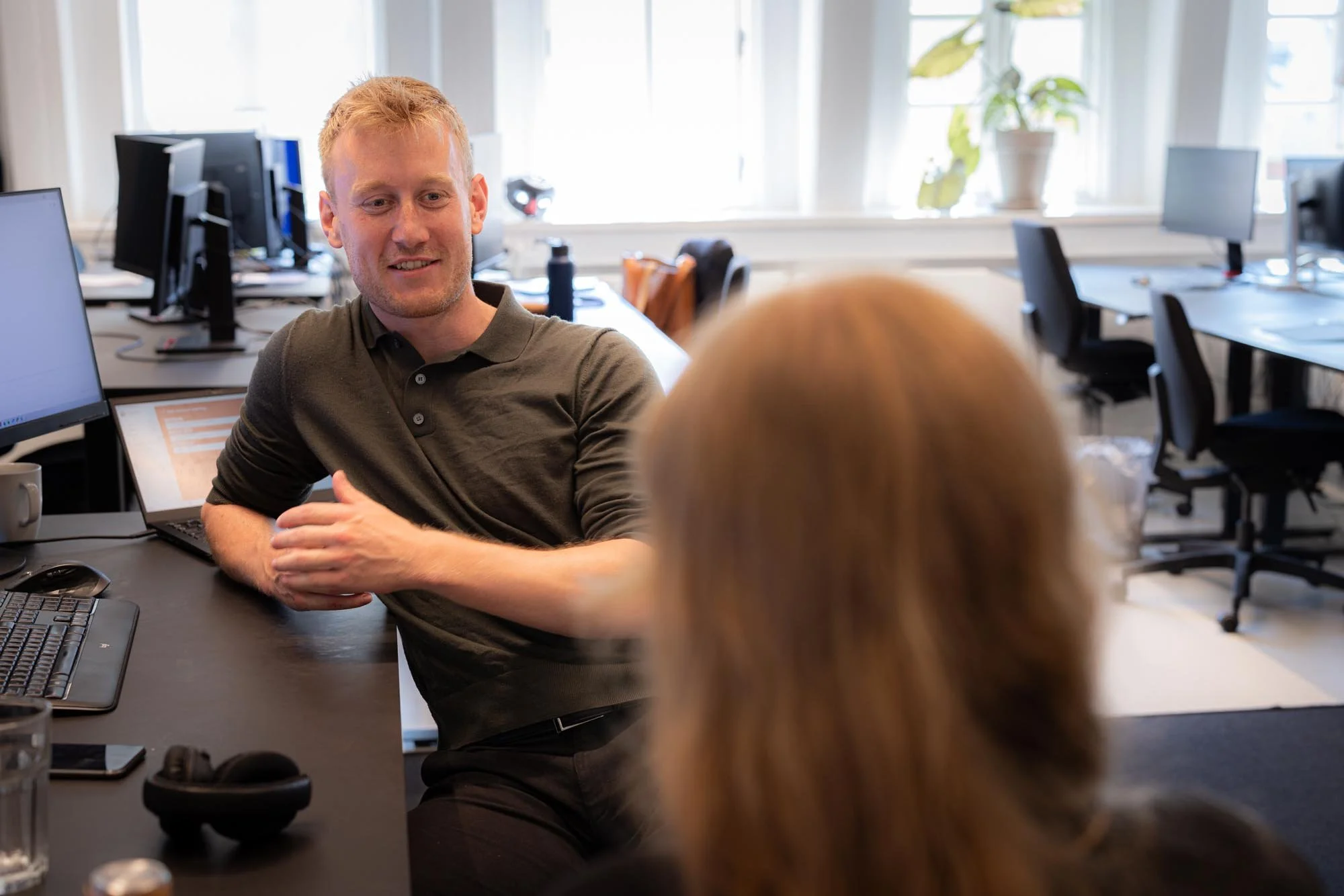 A man with short blond hair wearing a black polo shirt sitting at a desk having a conversation with a woman with long blonde hair in an office setting with multiple computer monitors and desks.