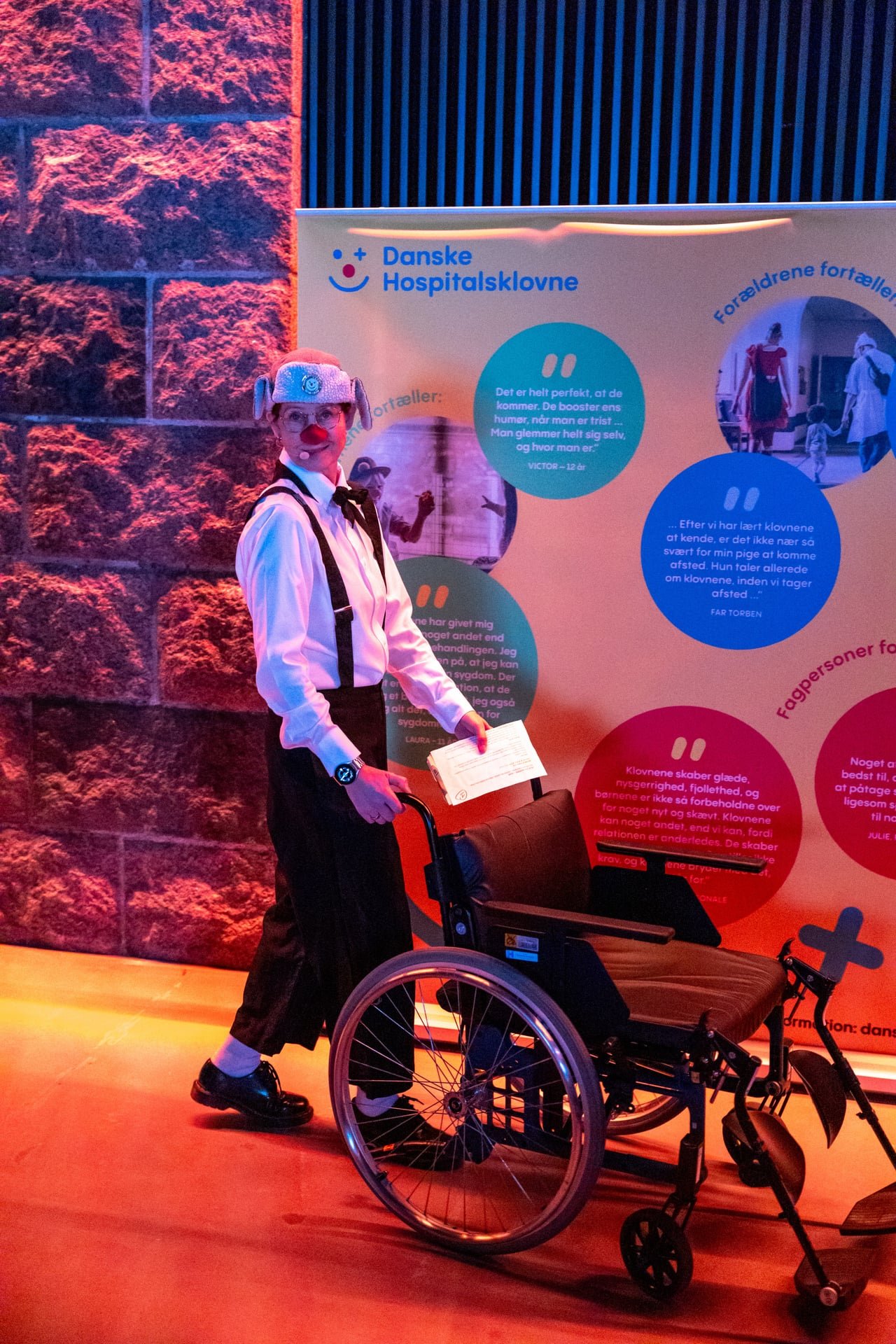 Person dressed as a clown with a wheelchair stands in front of a colorful informational display.