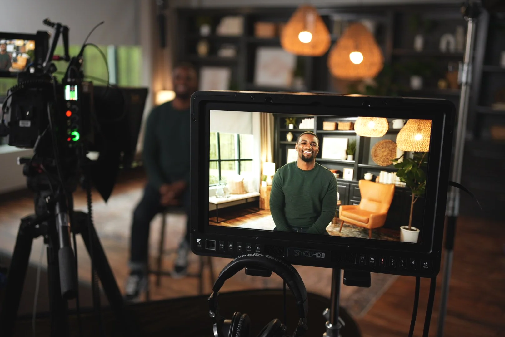 A man is sitting in a well-lit study room with a window, orange chair, and decorative wall shelves, smiling for a video recording seen on a camera monitor, with a camera and equipment in the foreground.