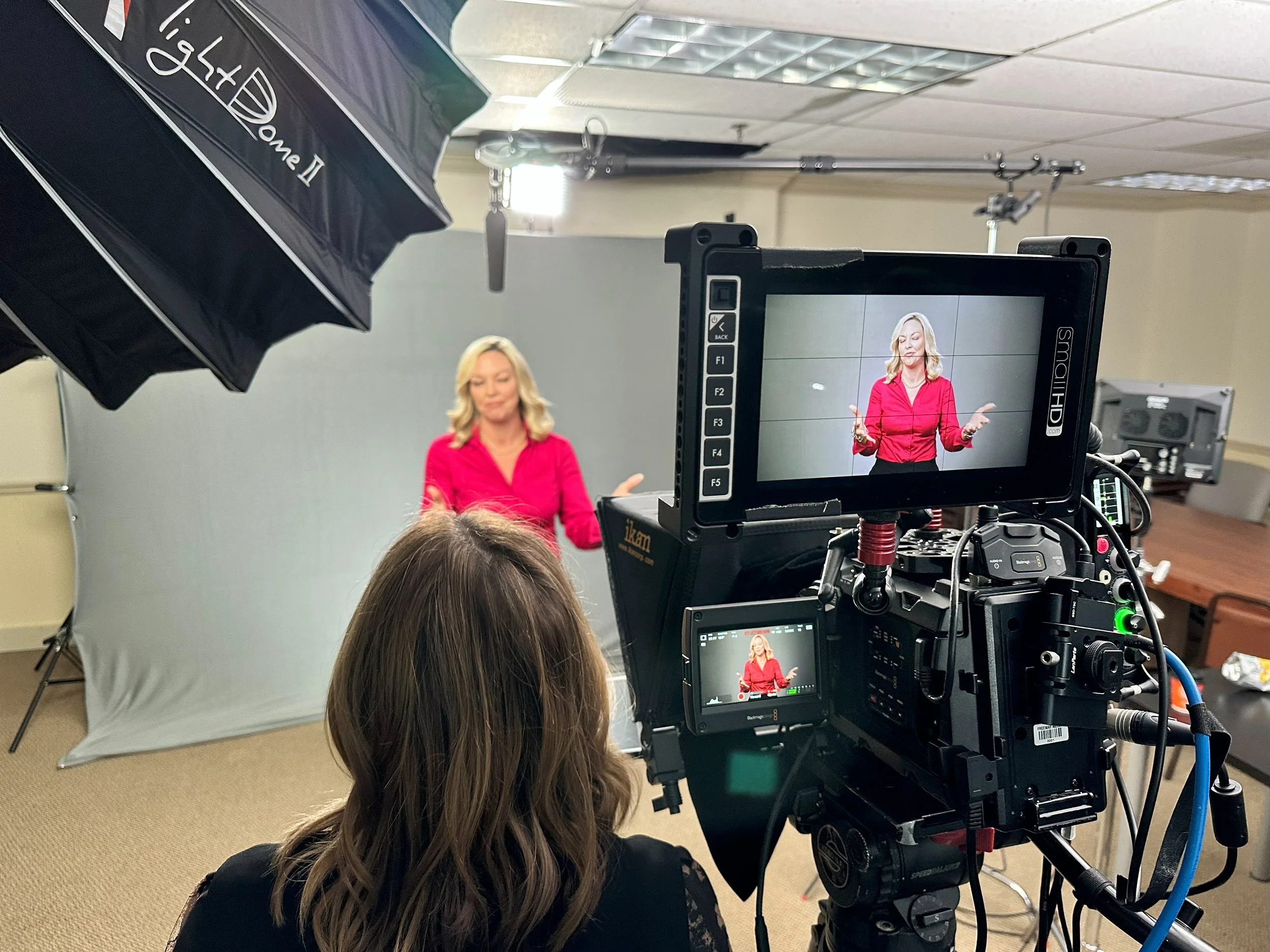 A behind-the-scenes view of a video production setup with a woman in a red blouse on camera, large lights, and video monitors showing her image.