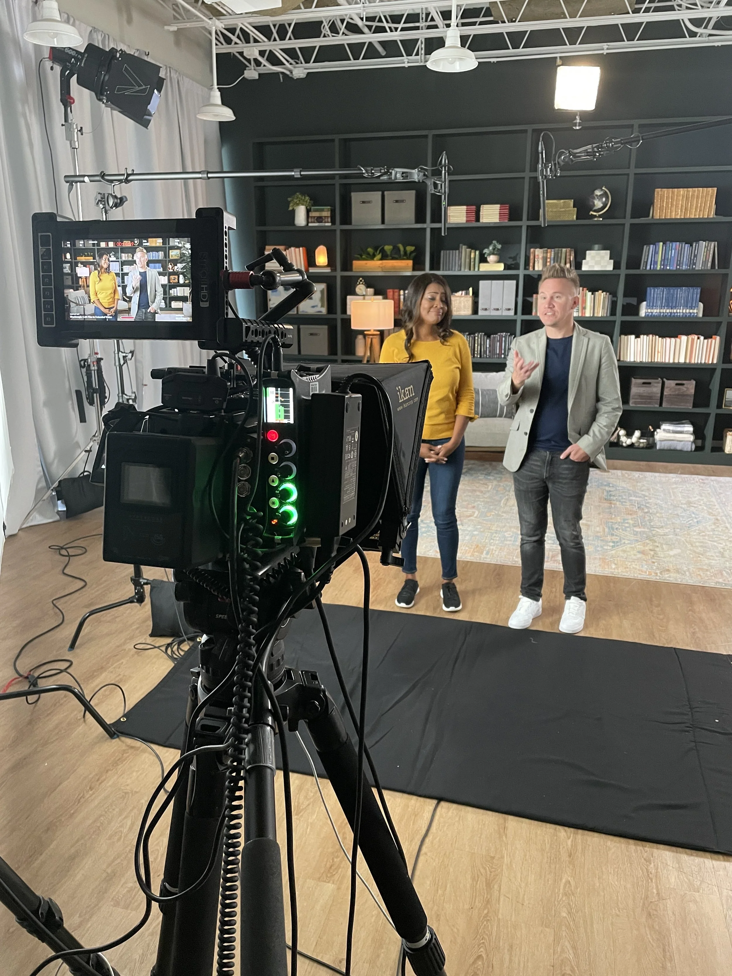 Two hosts, a woman in a yellow sweater and a man in a gray blazer, standing in front of a camera in a studio with a bookshelf background. The woman is looking at the man, who is speaking, while the camera captures the scene.
