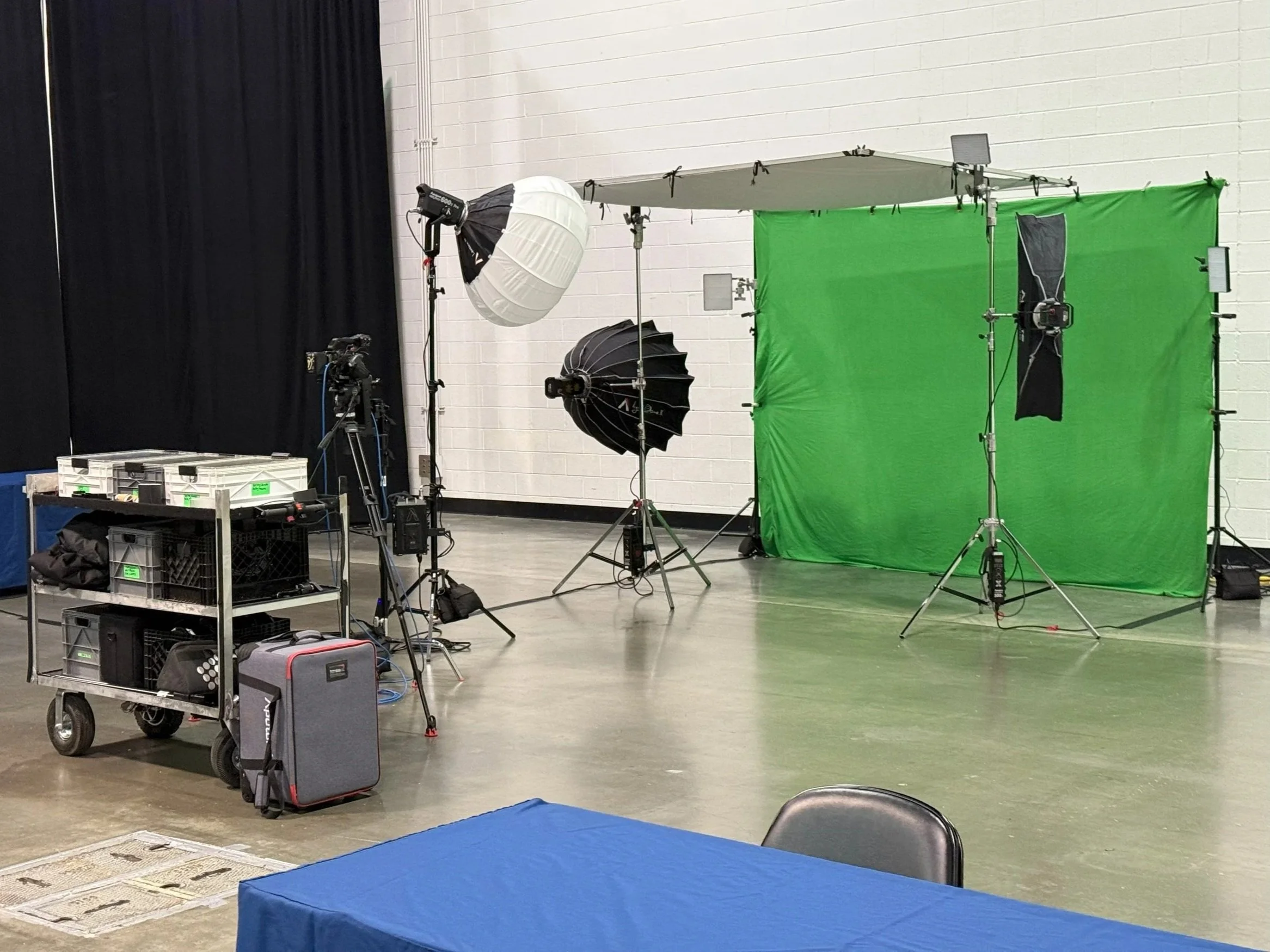 Photography studio setup with green screen, professional lighting, cameras, and equipment on a C-stand, with a blue table and black chairs in the foreground.