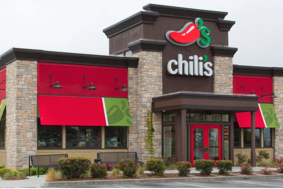 Exterior of a Chili's restaurant with stone and brick facade, red awnings, benches, and the Chili's logo with a red chili pepper sign