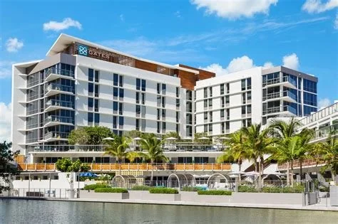 Modern multi-story hotel with glass balconies and palm trees in front, situated by the water under a blue sky.