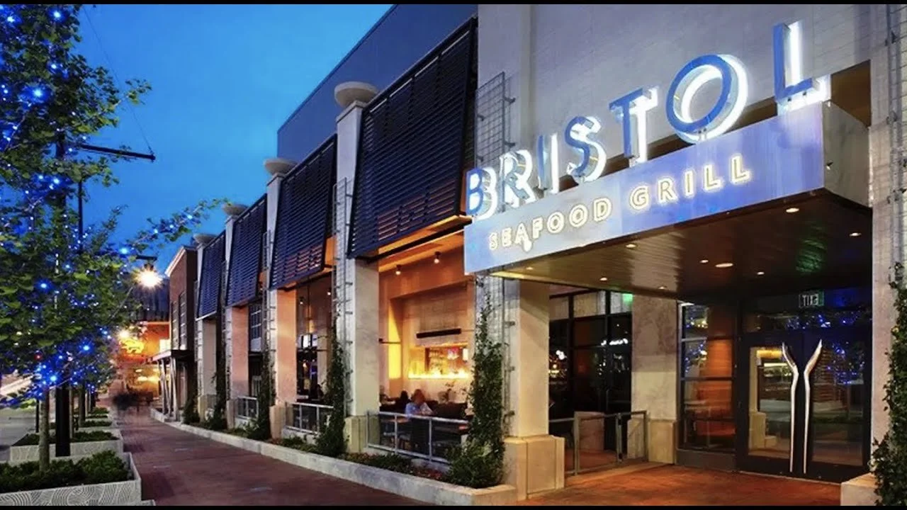 Exterior view of a seafood restaurant named 'BRISTOL'. The sign is illuminated and the restaurant has outdoor seating with people dining inside. There are trees with blue string lights on the sidewalk.