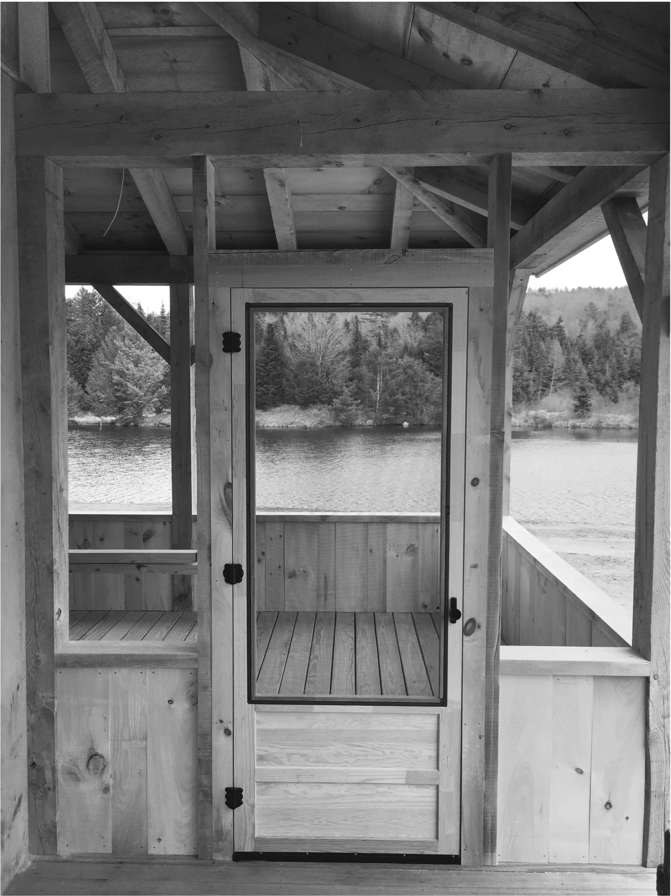 Black and white photo of the wooden interior of a small house or cabin under construction, featuring a door with a screen, overlooking a body of water and a forested shoreline outside. Built by the Timber, Stone, & Steel lead contractor, Taylor Prout