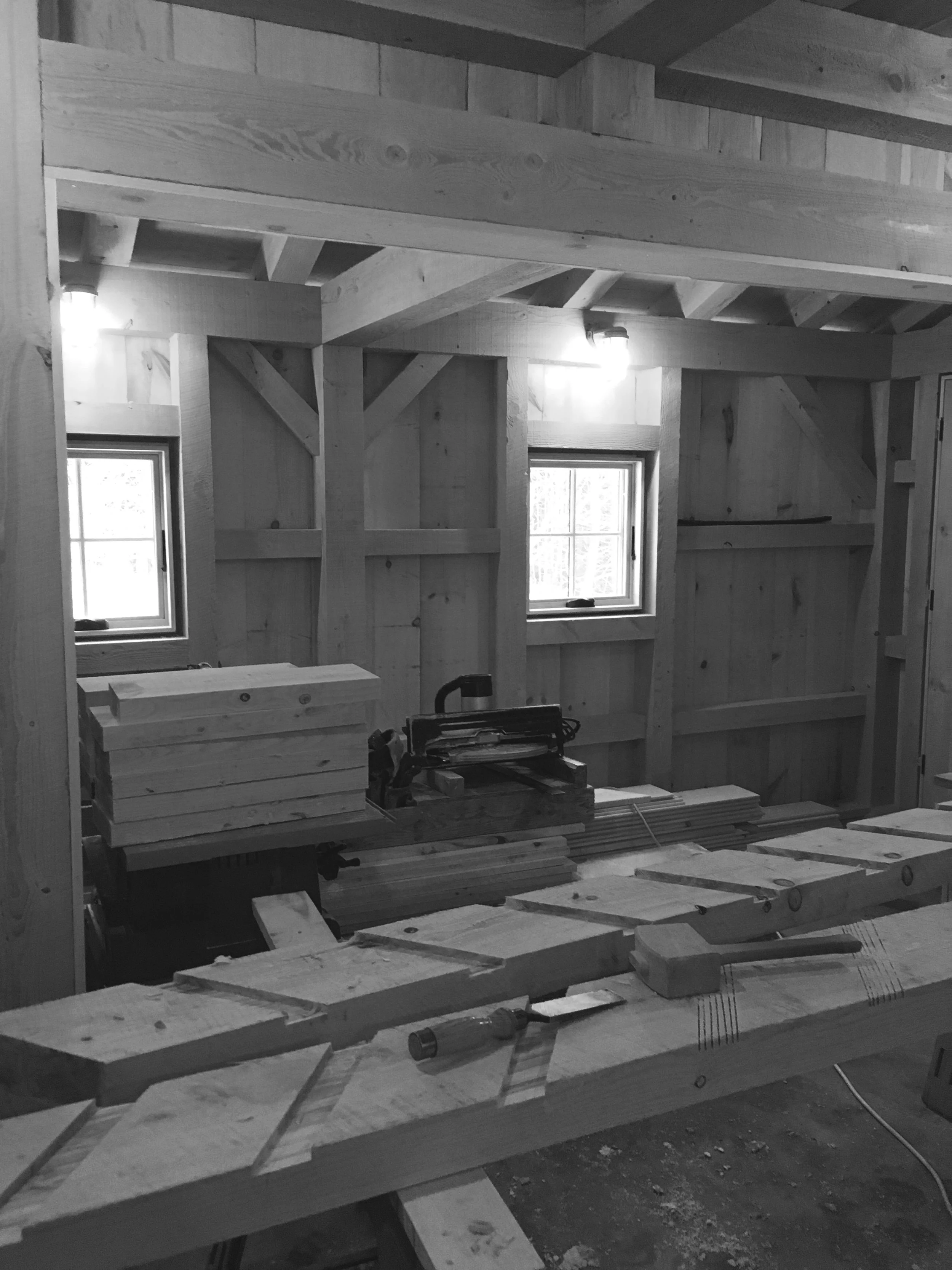 Interior view of a woodworking workshop with wooden beams, a workbench with wood pieces, tools, and two small windows. Built by the Timber, Stone, & Steel lead contractor, Taylor Prouty, in Vermont.