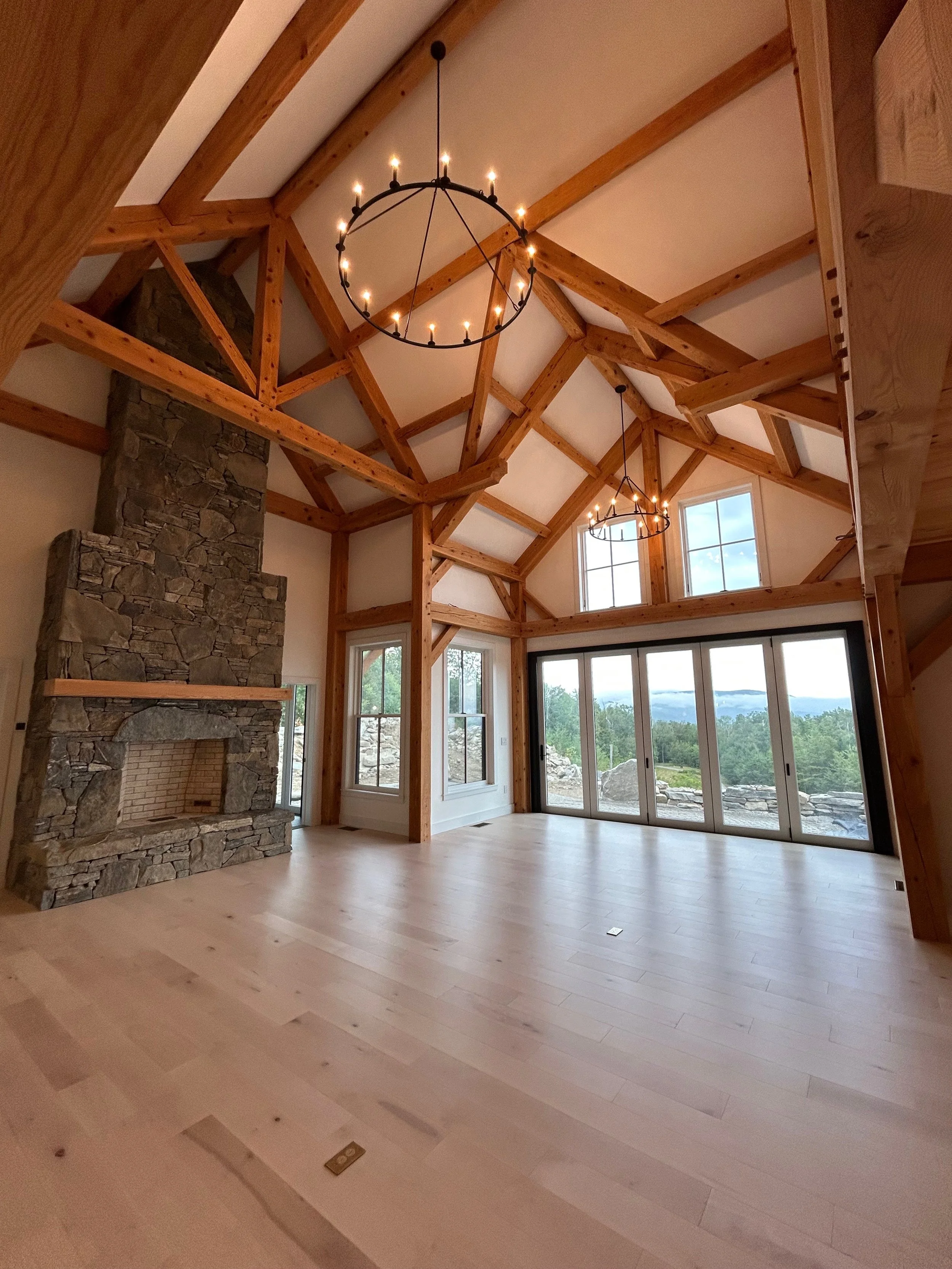 Empty living room with large glass sliding doors, stone fireplace, timberframe beams, and two circular chandeliers with candles, overlooking a scenic outdoor view. Built by the Timber, Stone, & Steel lead contractor, Taylor Prouty in Vermont.