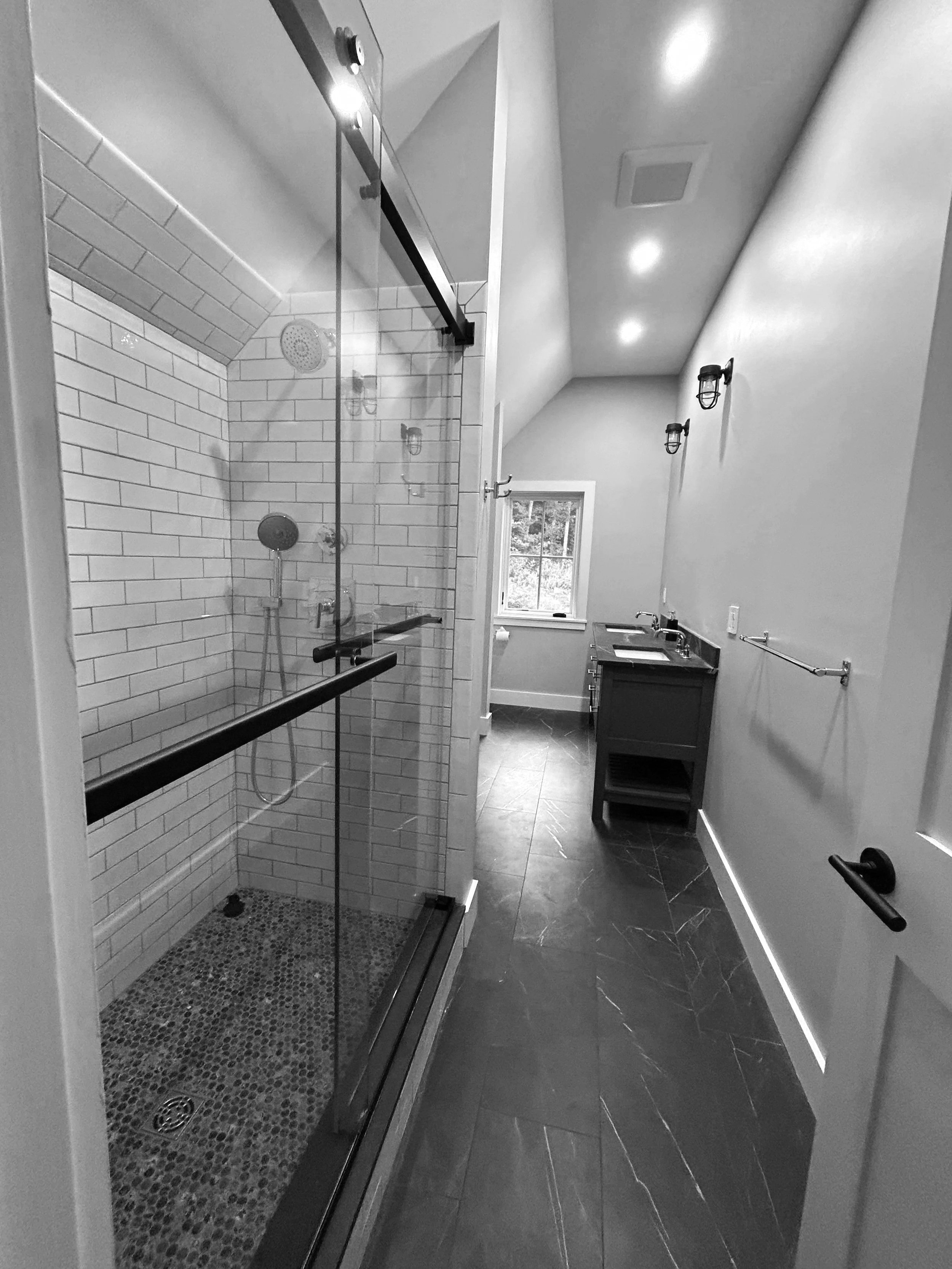 A modern bathroom with a walk-in shower with glass doors, white subway tile walls, pebble tile flooring, and a small vanity with a mirror and window in the background. Built by the Timber, Stone, & Steel lead contractor, Taylor Prouty in Vermont.