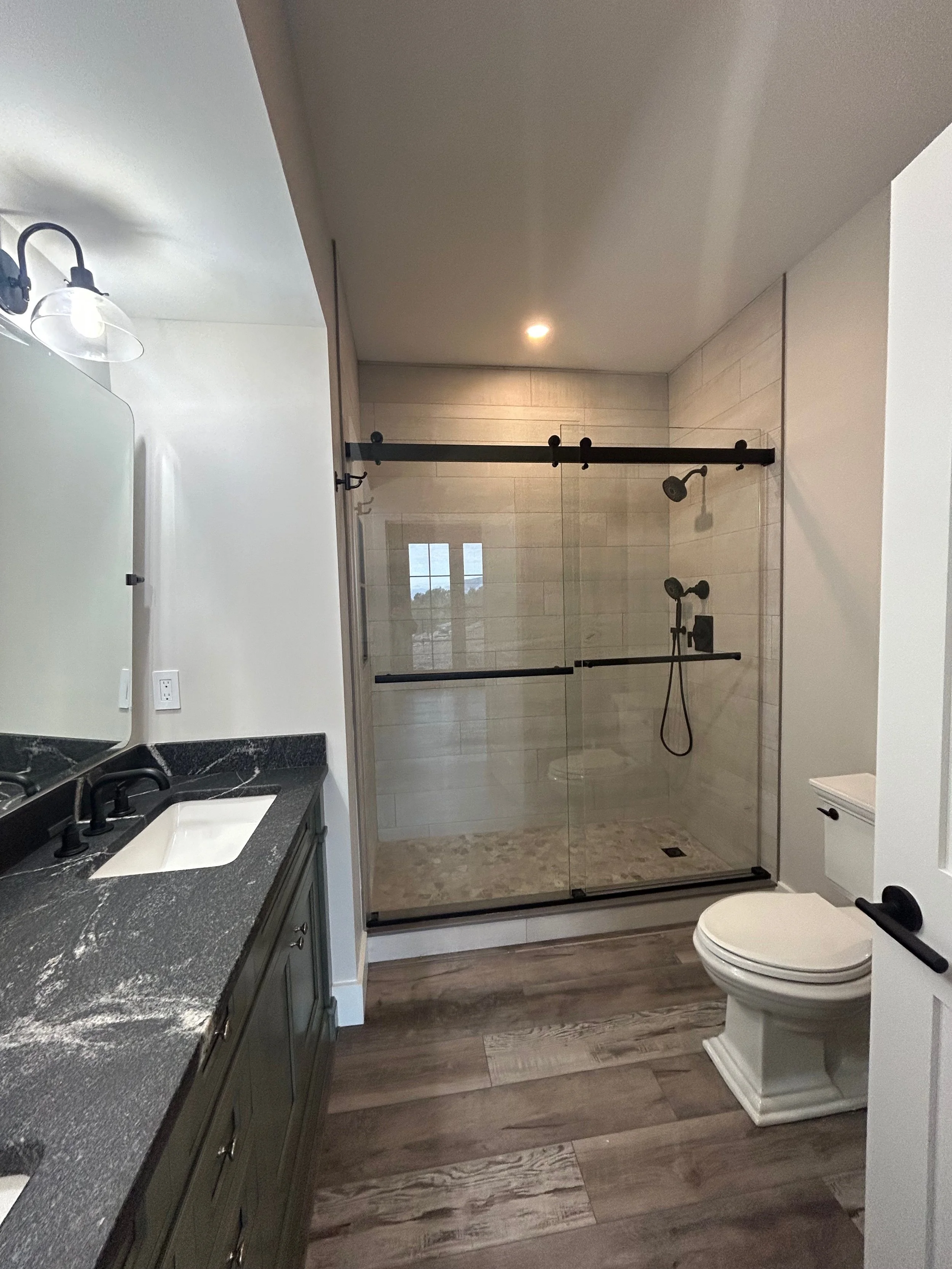 Modern bathroom with a double sink vanity, black fixtures, a walk-in glass shower with black hardware, and a white toilet, wooden flooring, and neutral wall colors. Built by the Timber, Stone, & Steel lead contractor, Taylor Prouty, in Vermont.