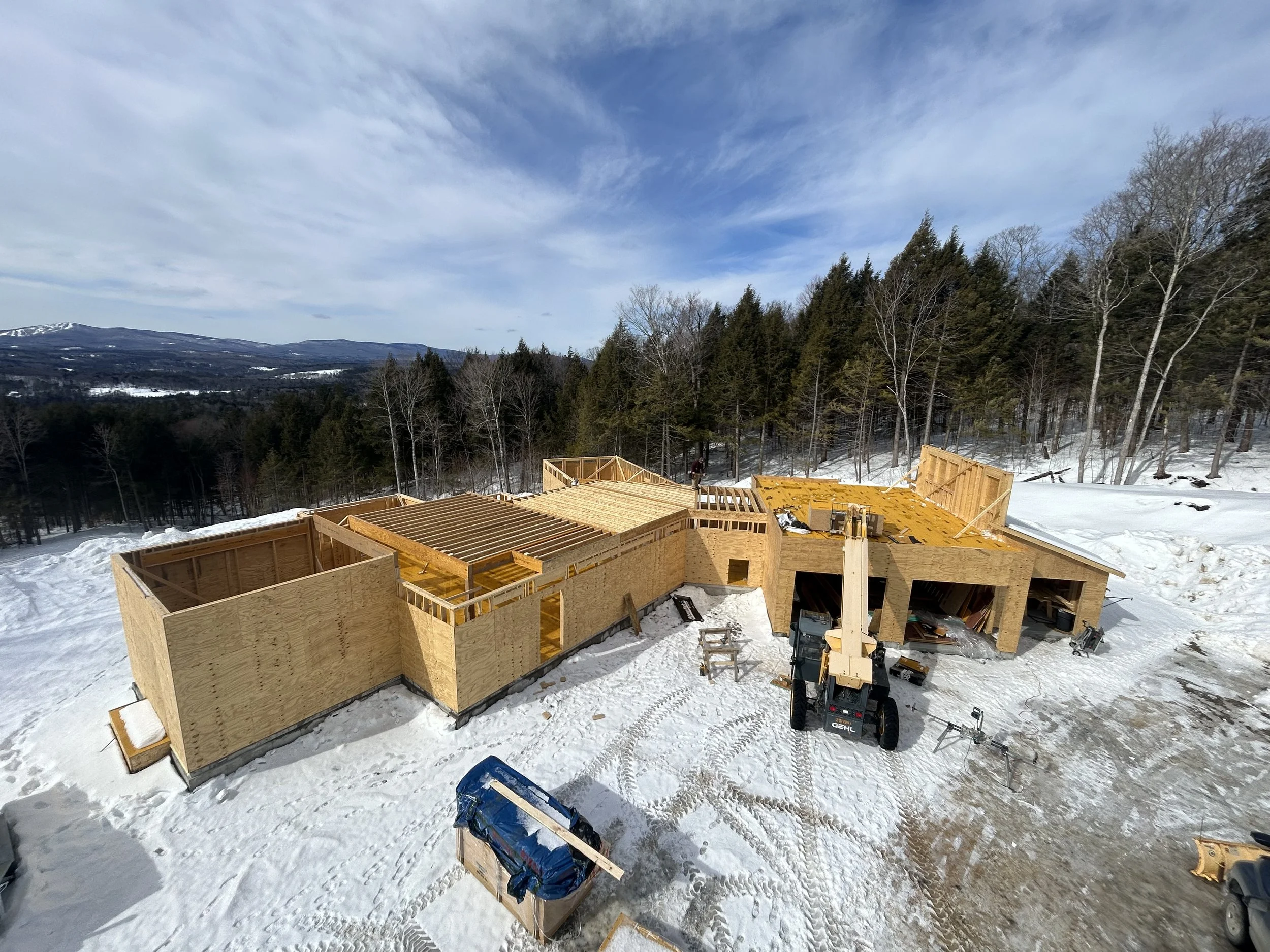 Snowy construction site with wooden framing for a house, surrounded by trees and mountains in the background. Built by Timber, Stone, & Steel, lead contractor Taylor Prouty in Vermont.