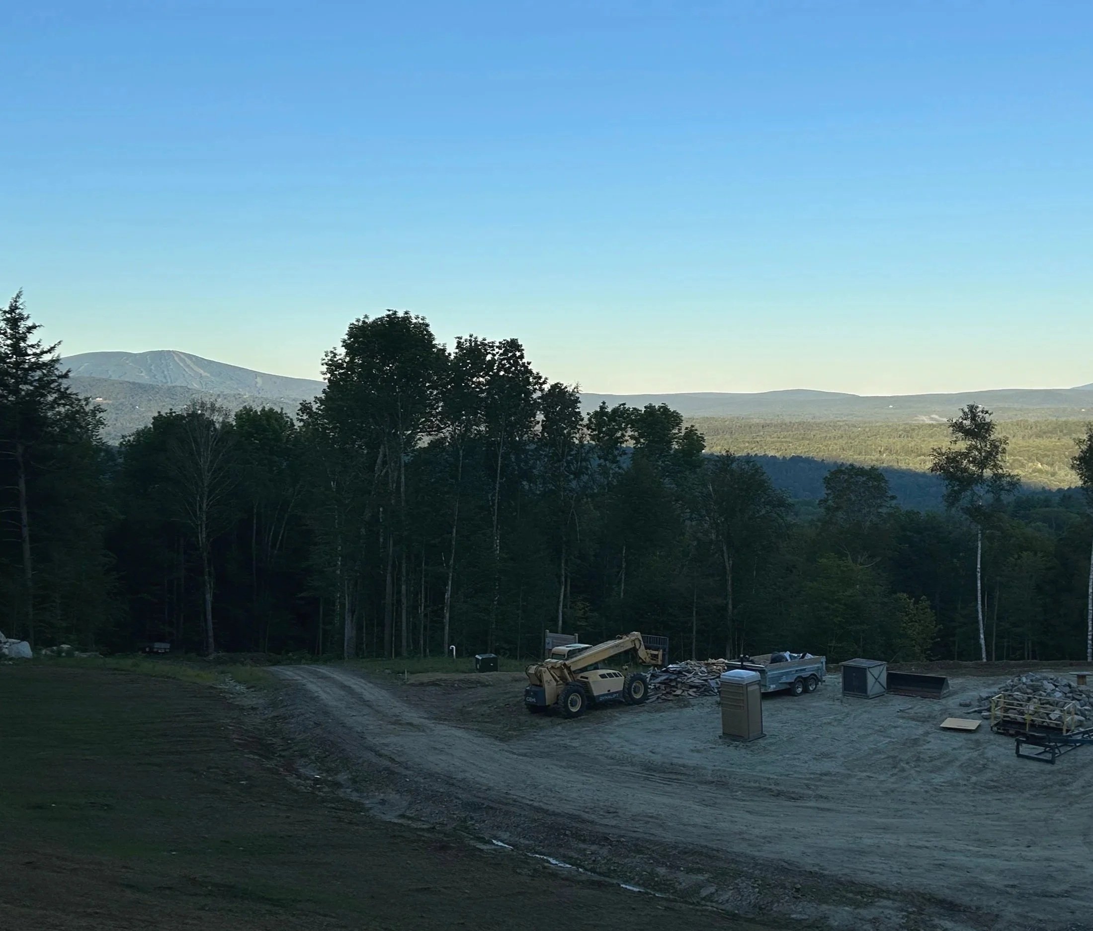 A construction site with a backhoe loader, a pickup truck, and construction materials set against a backdrop of forested hills and mountains under a clear blue sky. Built by the Timber, Stone, & Steel lead contractor, Taylor Prouty in Vermont.