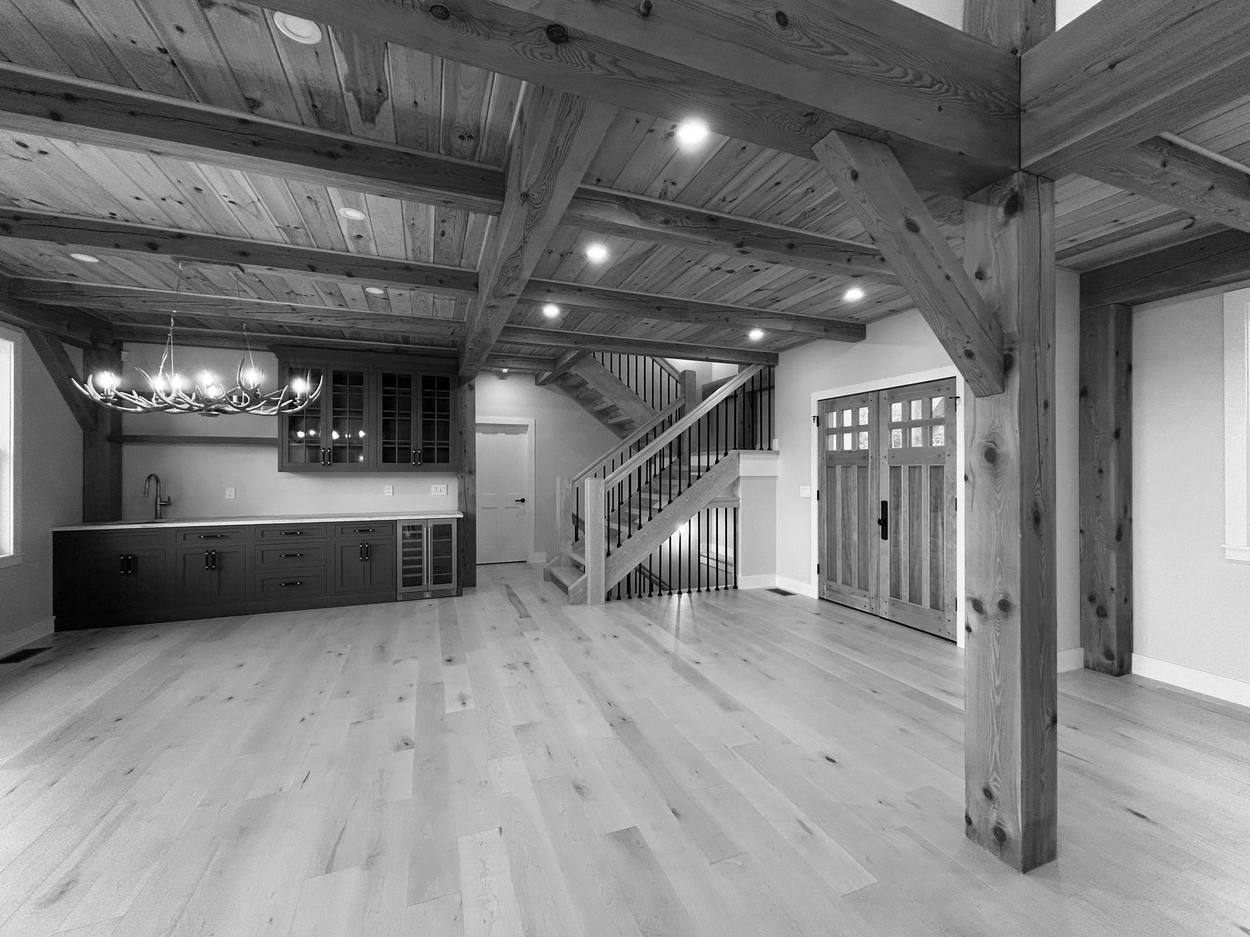 Interior of a rustic living space with a timber frame ceiling, hardwood floors, a staircase, wooden double doors, and a built-in bar area with cabinets and a chandelier. Built by the Timber, Stone, & Steel lead contractor, Taylor Prouty, in Vermont.