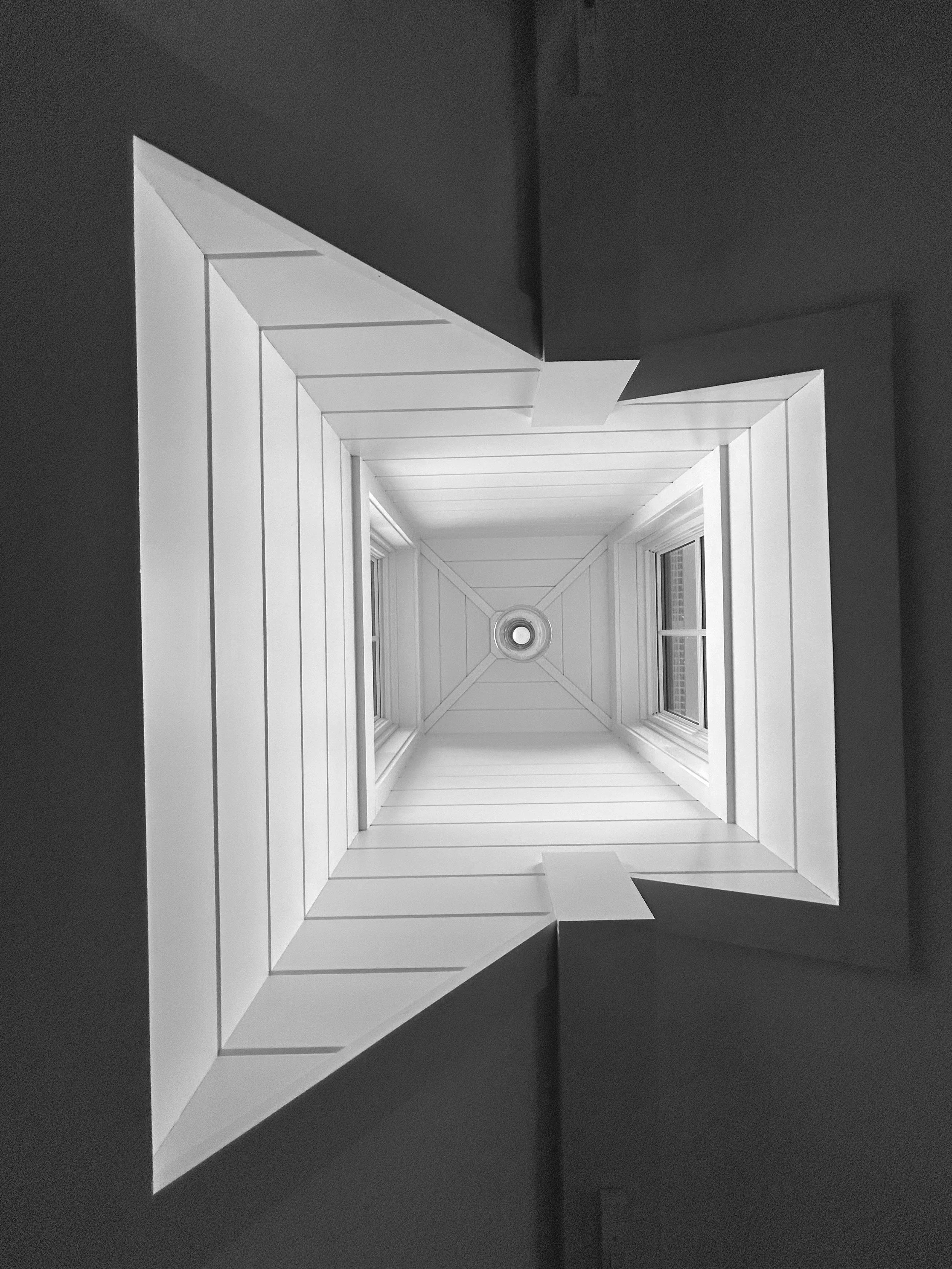A view looking up from the bottom of a square skylight with white trim and two windows, creating a geometric pattern in black and white. Built by the Timber, Stone, & Steel lead contractor, Taylor Prouty in Vermont.