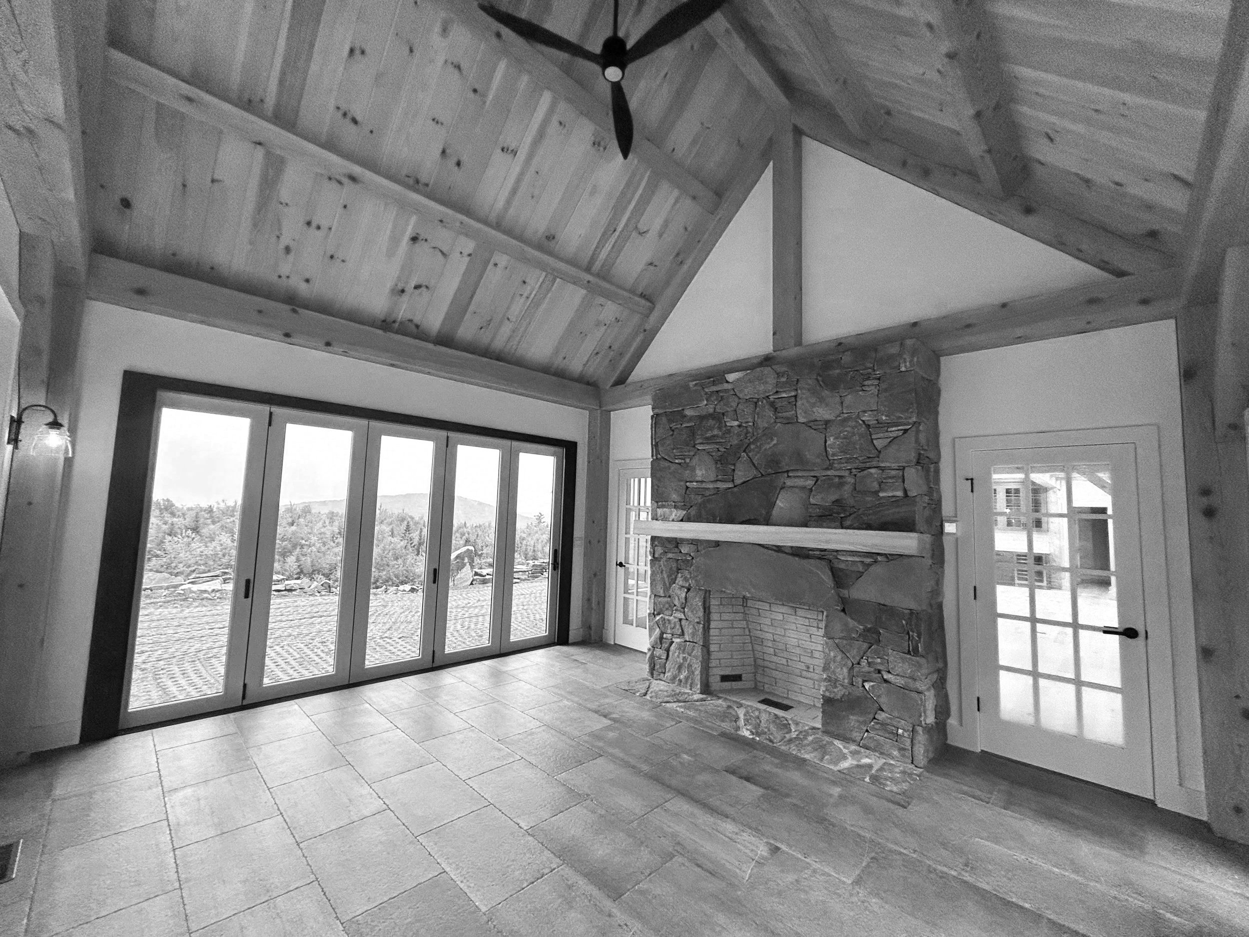 Interior view of a room with a vaulted timber frame ceiling, a stone fireplace, large sliding glass door, and a glass-paneled door leading outside, with tile flooring. Built by the Timber, Stone, & Steel lead contractor, Taylor Prouty in Vermont.
