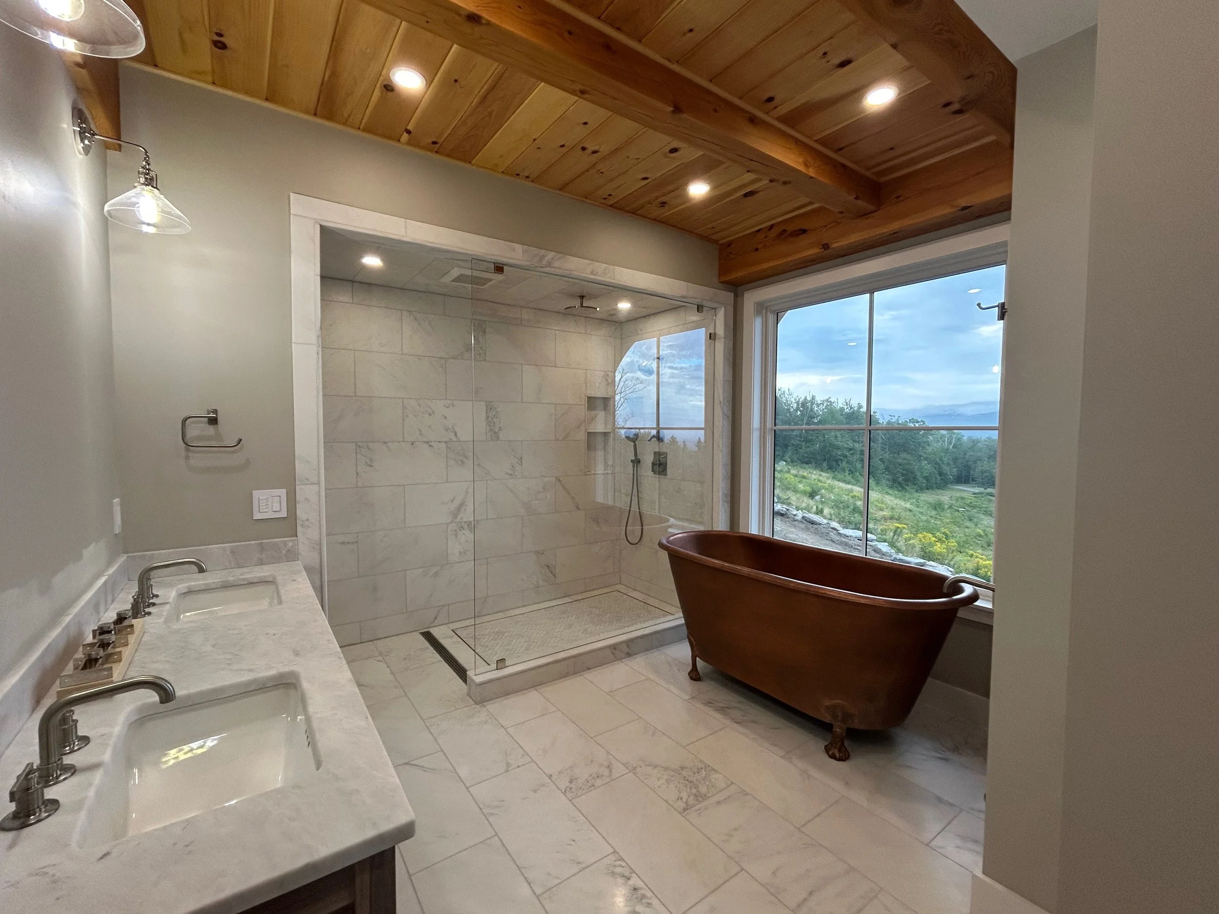 Bathroom with double vanity sinks, marble countertops, a glass-enclosed shower, a clawfoot bathtub, a large window with a scenic outdoor view, and a ceiling with recessed lighting. Built by the Timber, Stone, & Steel lead contractor, Taylor Prouty.