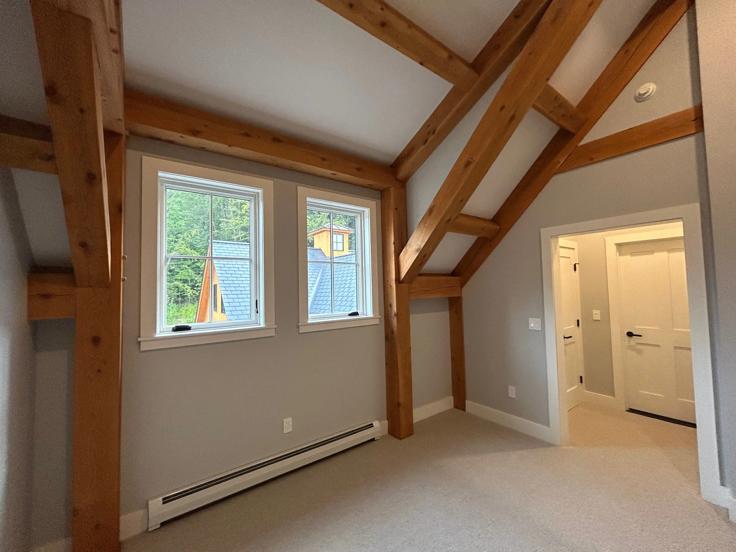 Empty room with light gray walls, beige carpet, wooden beams, and two windows showing a house and trees outside. Built by the Timber, Stone, & Steel lead contractor, Taylor Prouty, in Vermont.