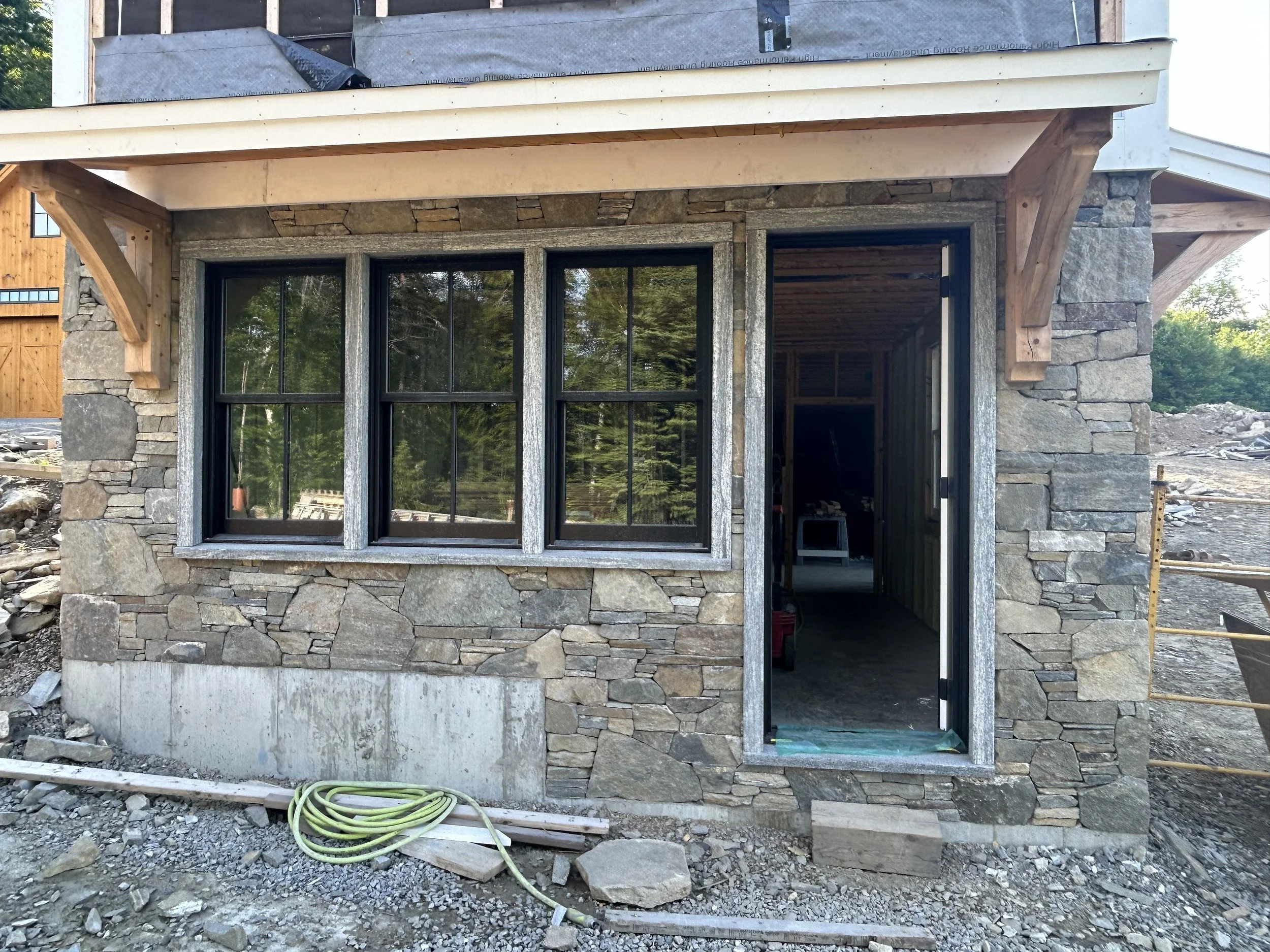 Construction site of a house with stonework exterior walls, three large window panes, and an open doorway. Building materials and tools are visible around the site. Built by Timber, Stone, & Steel, lead contractor Taylor Prouty in Vermont.