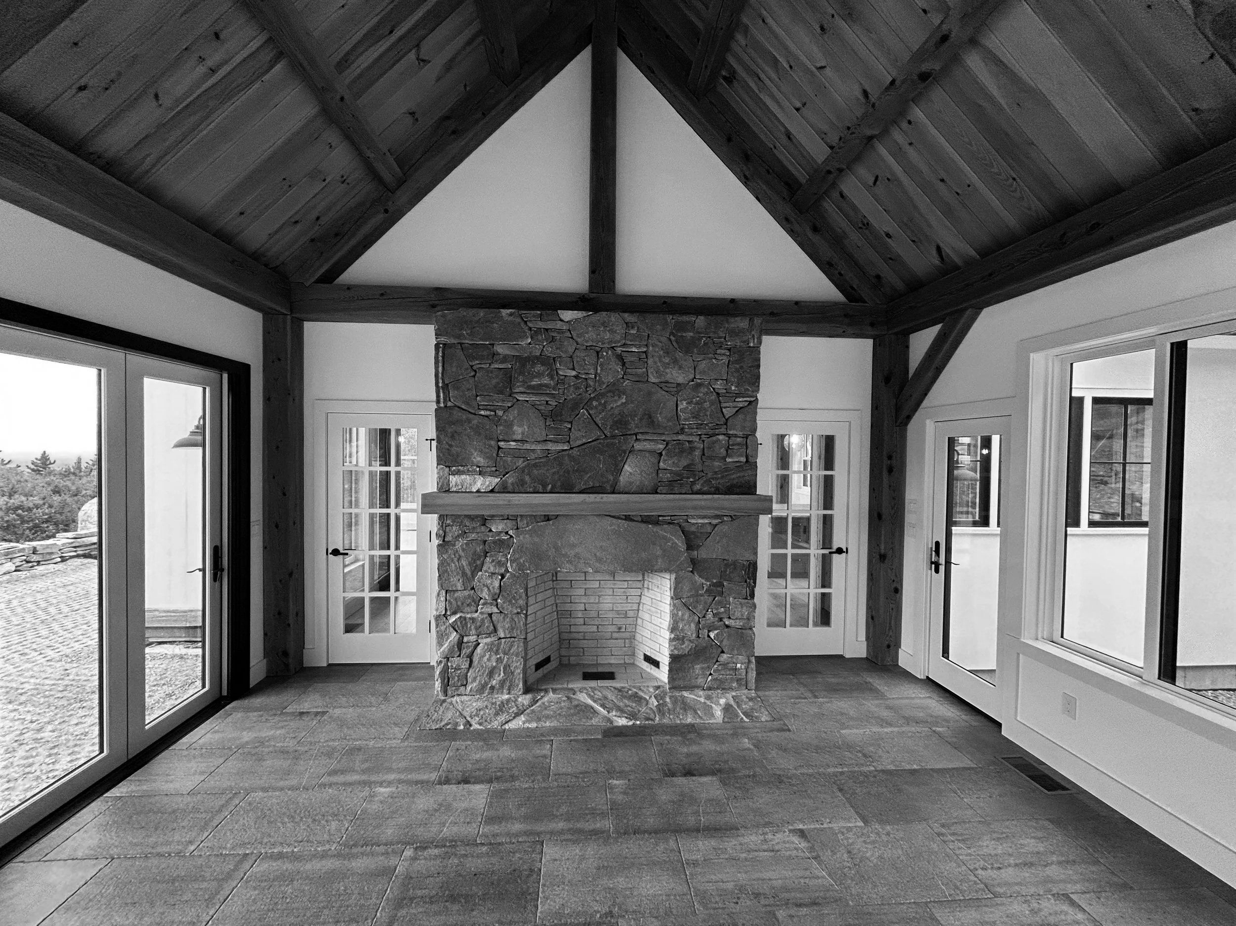 Living room with a stone fireplace, wooden vaulted ceiling, and large windows and glass doors with views outside. Built by the Timber, Stone, & Steel lead contractor, Taylor Prouty in Vermont.