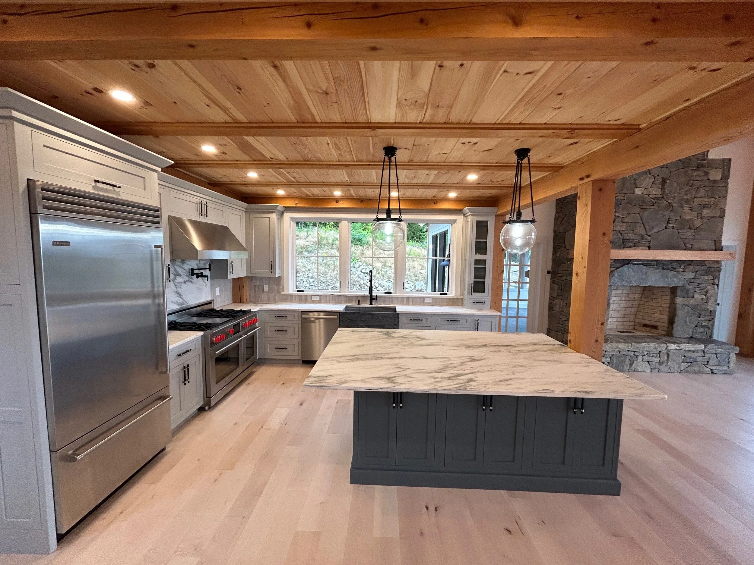 A modern kitchen with white cabinets, stainless steel appliances, a large marble island, and a wood ceiling. It features a stone fireplace and large windows at the back. Built by the Timber, Stone, & Steel lead contractor, in Vermont.