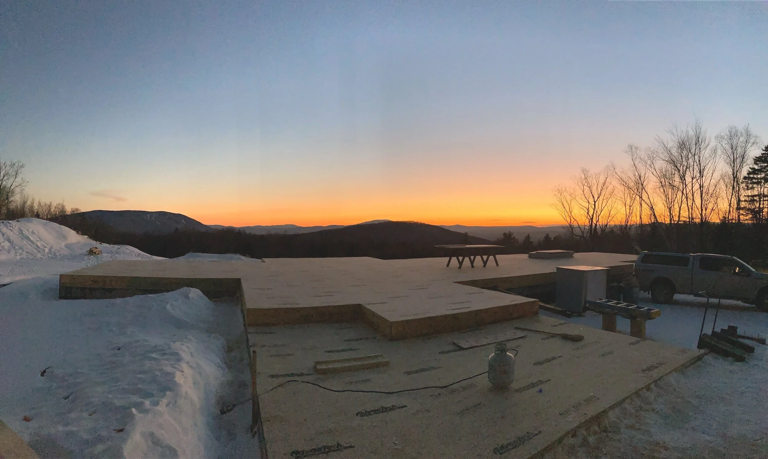 Construction site on a snowy landscape during sunset with a partially built wooden platform, a vehicle, and tools. Built by the Timber, Stone, & Steel lead contractor, Taylor Prouty, in Vermont.