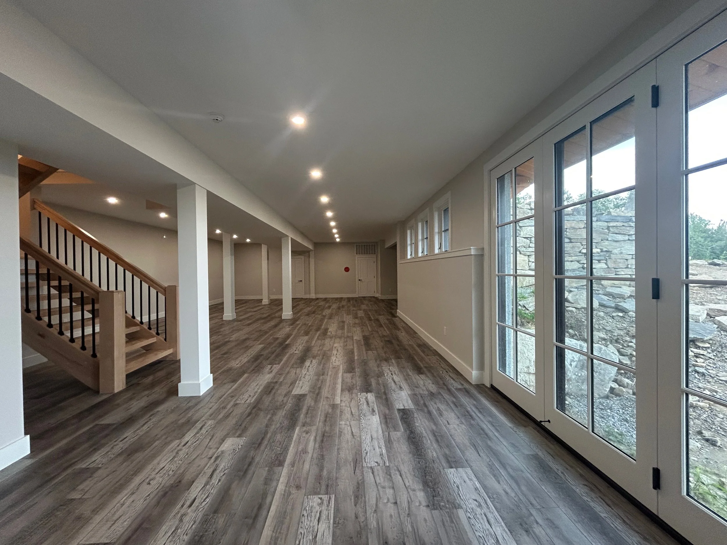 Empty spacious room with wood flooring, white walls, large glass doors, and a staircase leading upstairs. Built by the Timber, Stone, & Steel lead contractor, Taylor Prouty in Vermont.