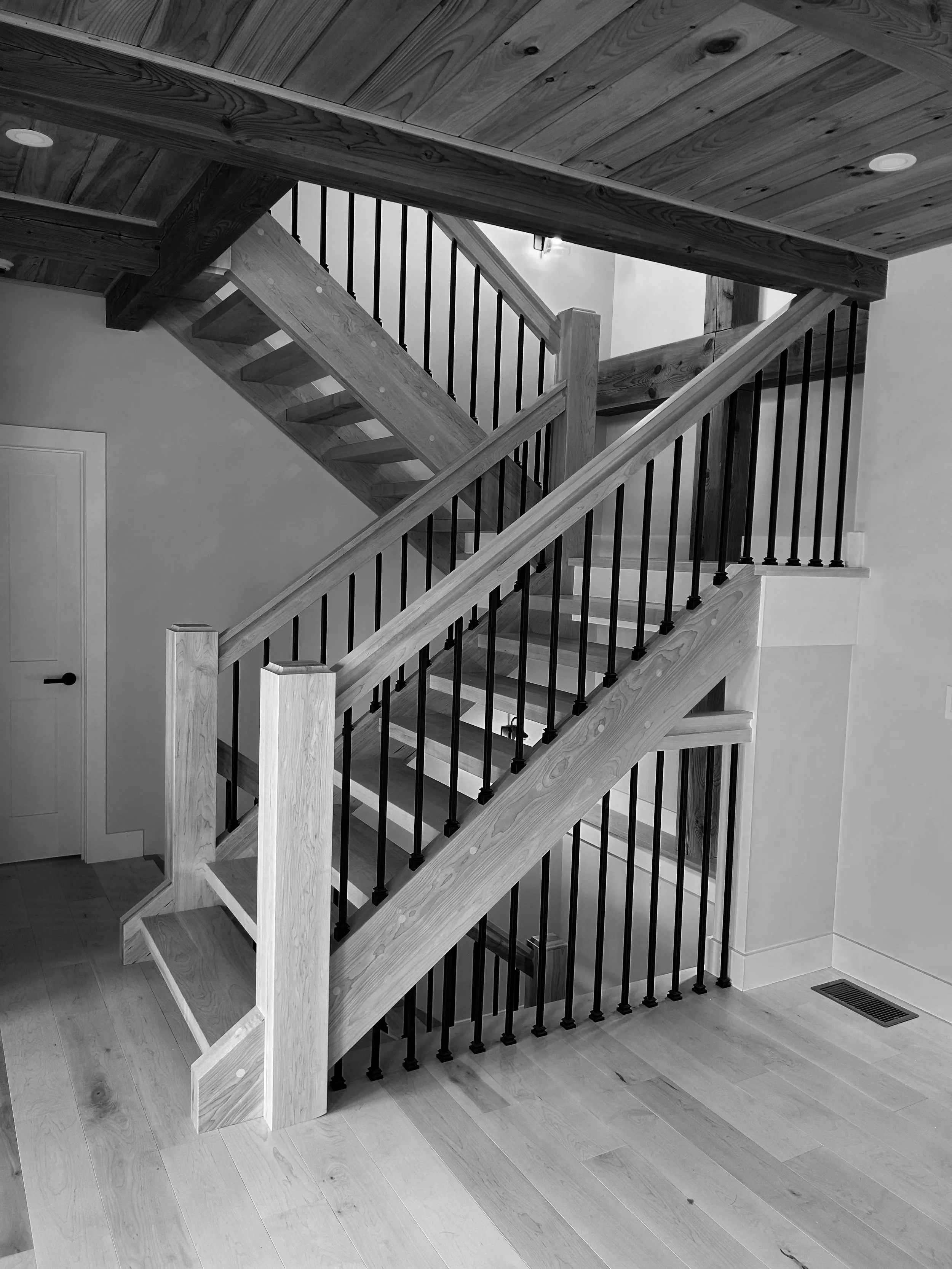 Black and white photo of a modern wooden staircase with black metal balusters, inside a house with hardwood floors and a ceiling with wooden paneling. Built by the Timber, Stone, & Steel lead contractor, Taylor Prouty in Vermont.