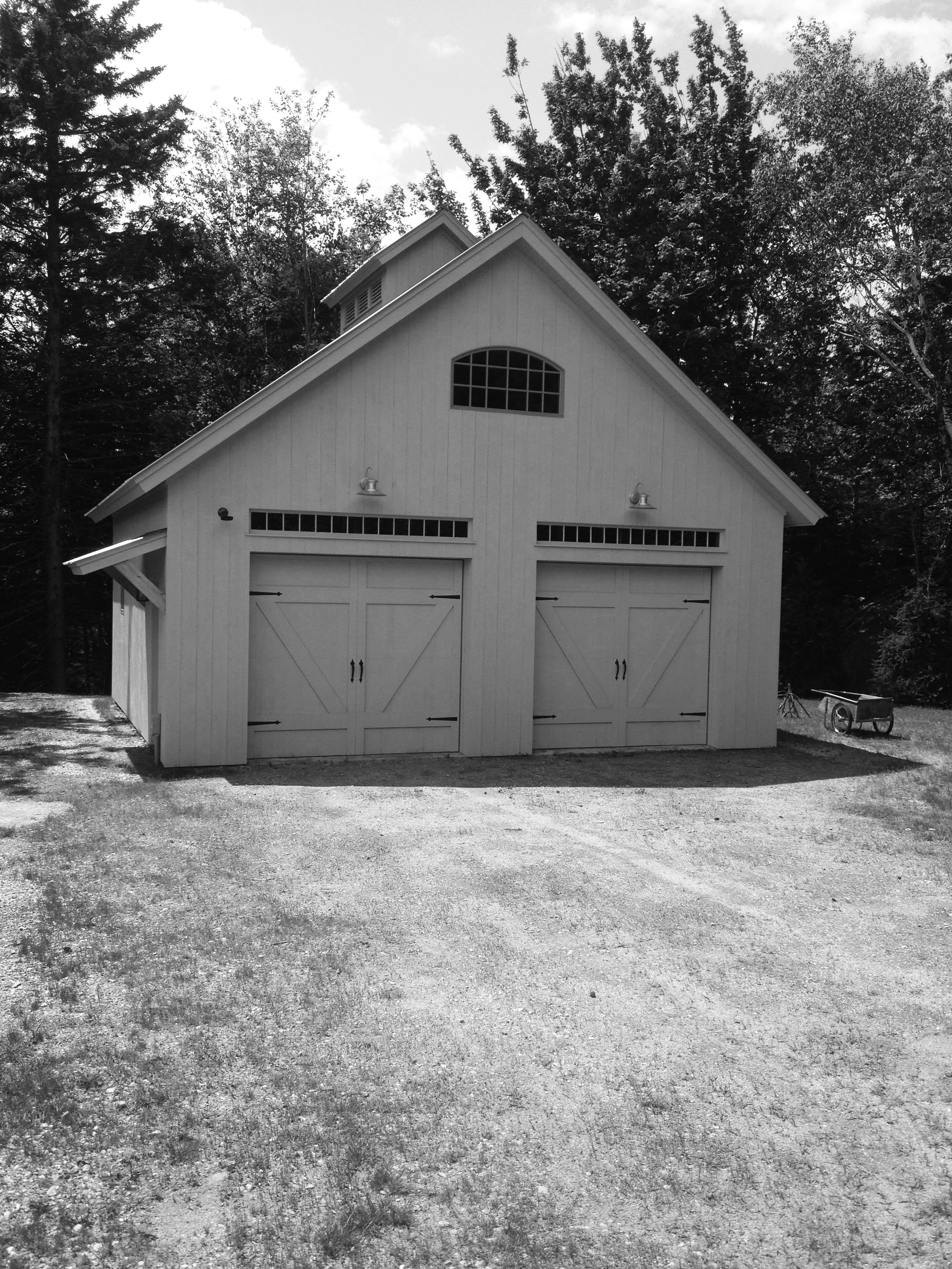 Large garage or barn with double doors, a small arched window above, and two wall-mounted lights. A gravel driveway. Built by the Timber, Stone, & Steel lead contractor, Taylor Prouty, in Vermont.