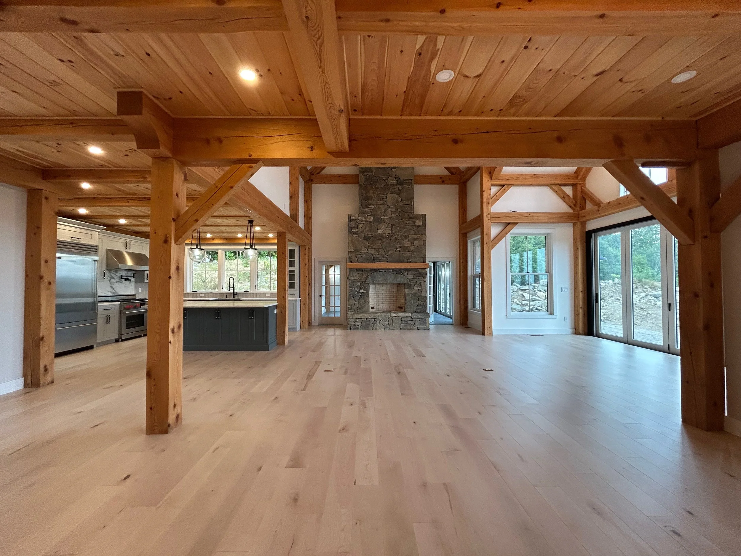 Open living room with wooden beams, stone fireplace, large windows, and sliding glass doors leading outside. Built by the Timber, Stone, & Steel lead contractor, Taylor Prouty in Vermont.