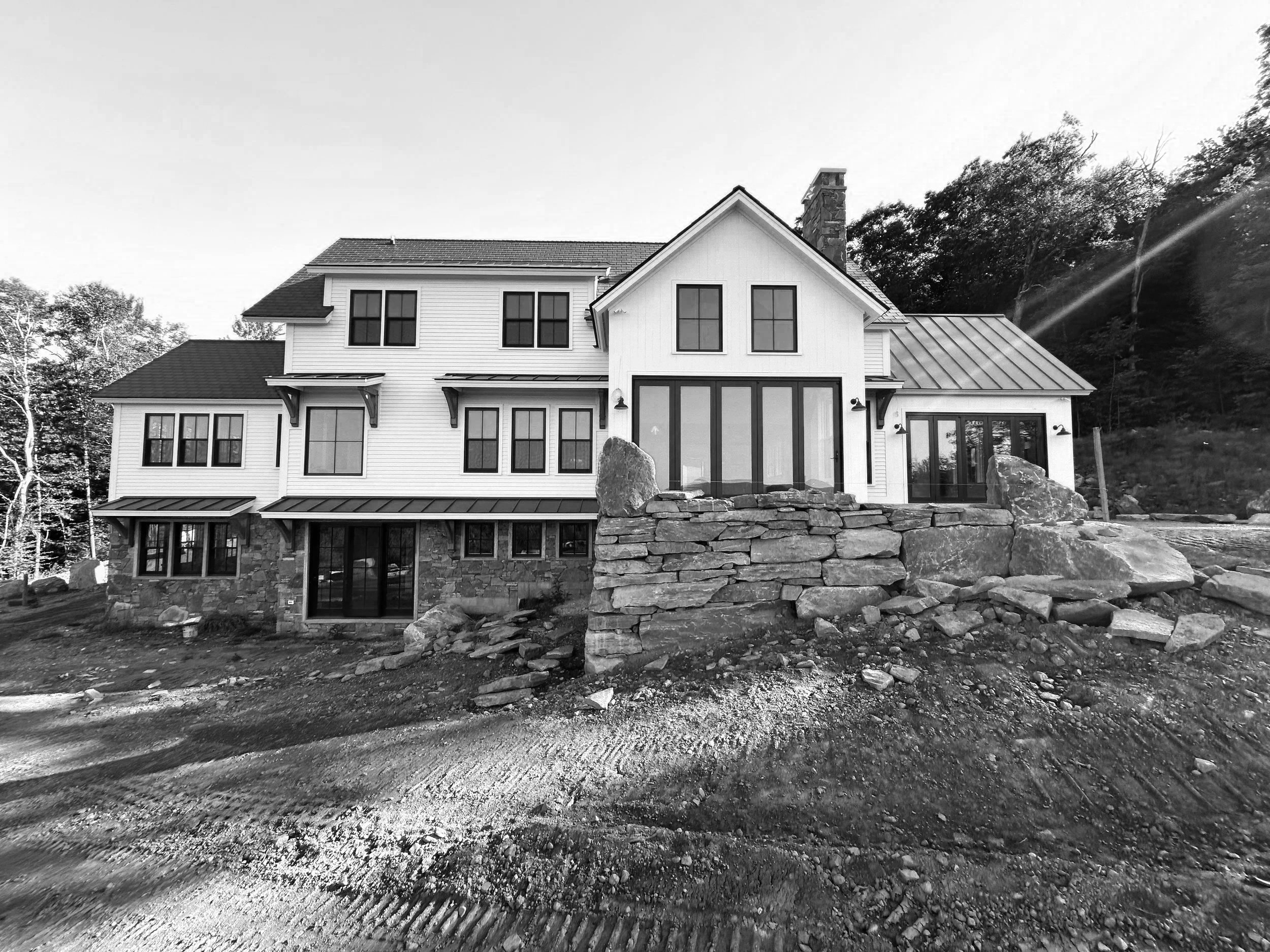 A large modern house under construction with a stone foundation, multiple windows, and a metal roof, set on uneven ground with construction debris. Built by the Timber, Stone, & Steel lead contractor, Taylor Prouty, in Vermont.