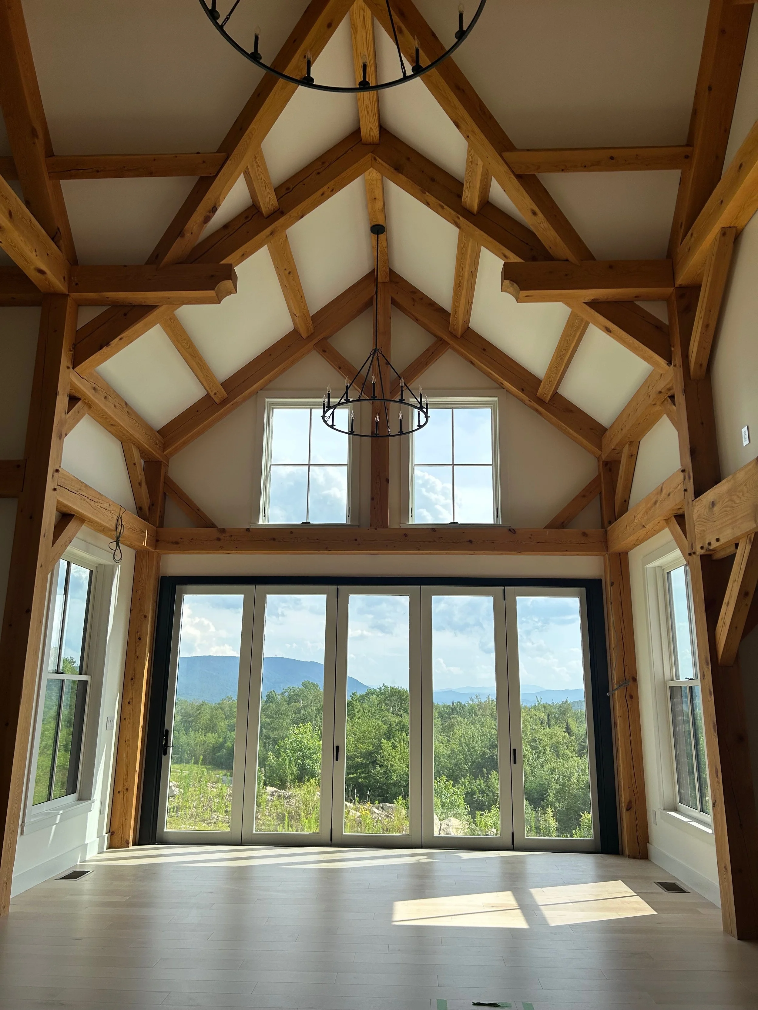 Interior view of a room with large glass doors and windows, timber frame ceiling, and a chandelier, showcasing a scenic mountain landscape outside. Built by the Timber, Stone, & Steel lead contractor, Taylor Prouty in Vermont.