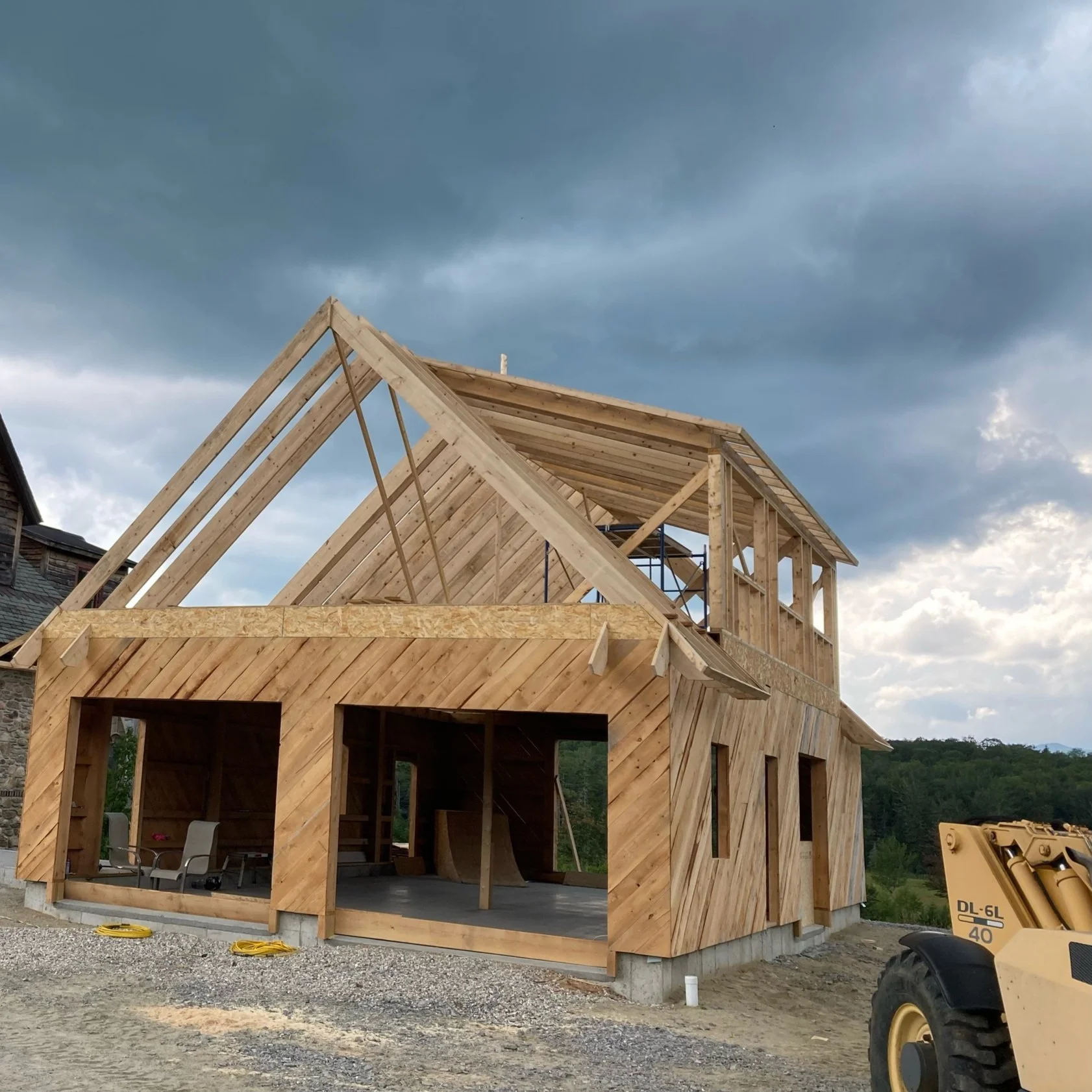 Timber frame house under construction with framing and partially completed walls, on a gravel lot with construction equipment nearby, and a cloudy sky overhead. Built by the Timber, Stone, & Steel lead contractor, Taylor Prouty, in Vermont.