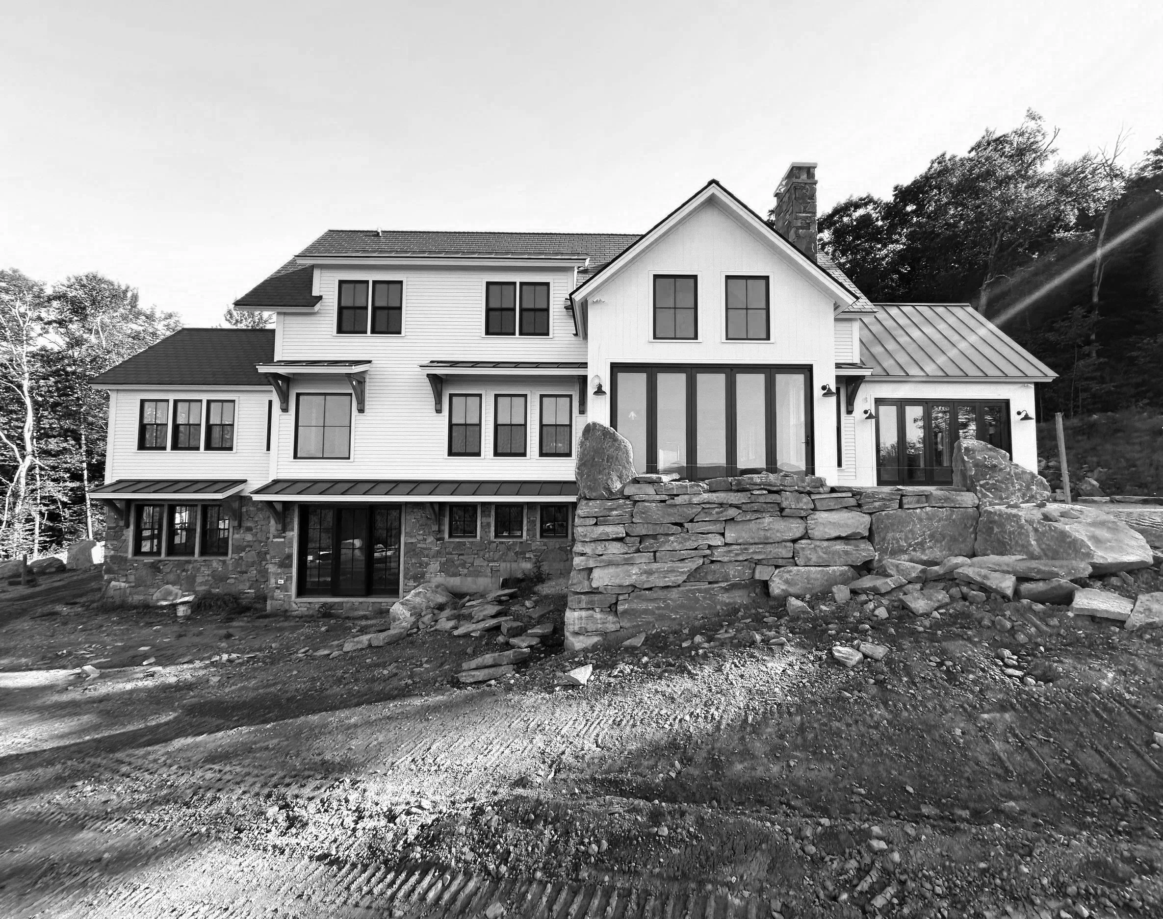 A large, multi-story house with a white exterior and black window frames, situated on a hill with a stone retaining wall and construction dirt in the foreground. Built by the Timber, Stone, & Steel lead contractor, Taylor Prouty, in Vermont.