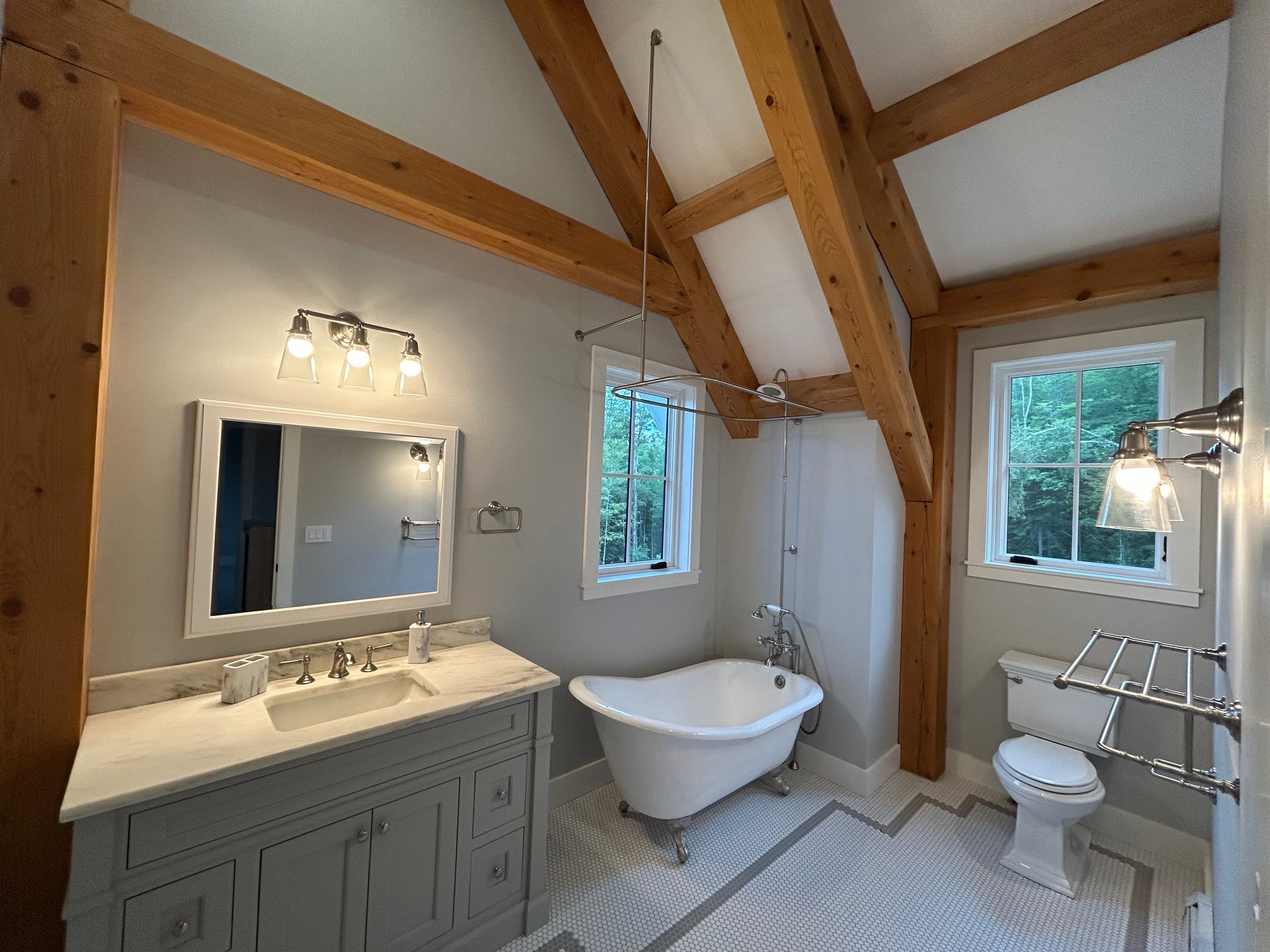 Bathroom with a timber frame ceiling, a clawfoot bathtub, a vanity with a marble countertop and mirror, a toilet, and two windows showing greenery outside. Built by the Timber, Stone, & Steel lead contractor, Taylor Prouty in Vermont.