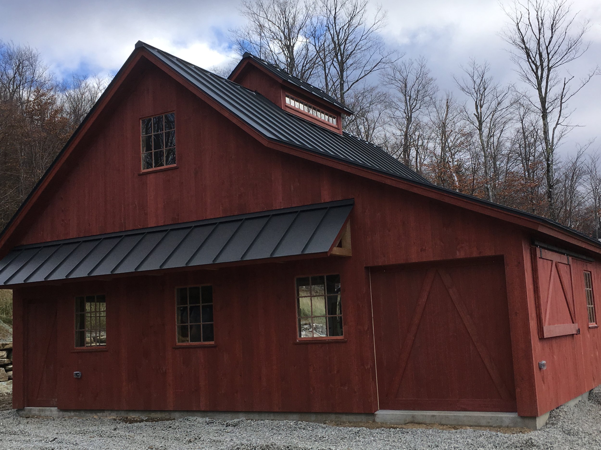Red barn-style building with black metal roof, small windows, and wooden sliding barn door, set against a background of leafless trees and cloudy sky. Built by the Timber, Stone, & Steel lead contractor, Taylor Prouty, in Vermont.