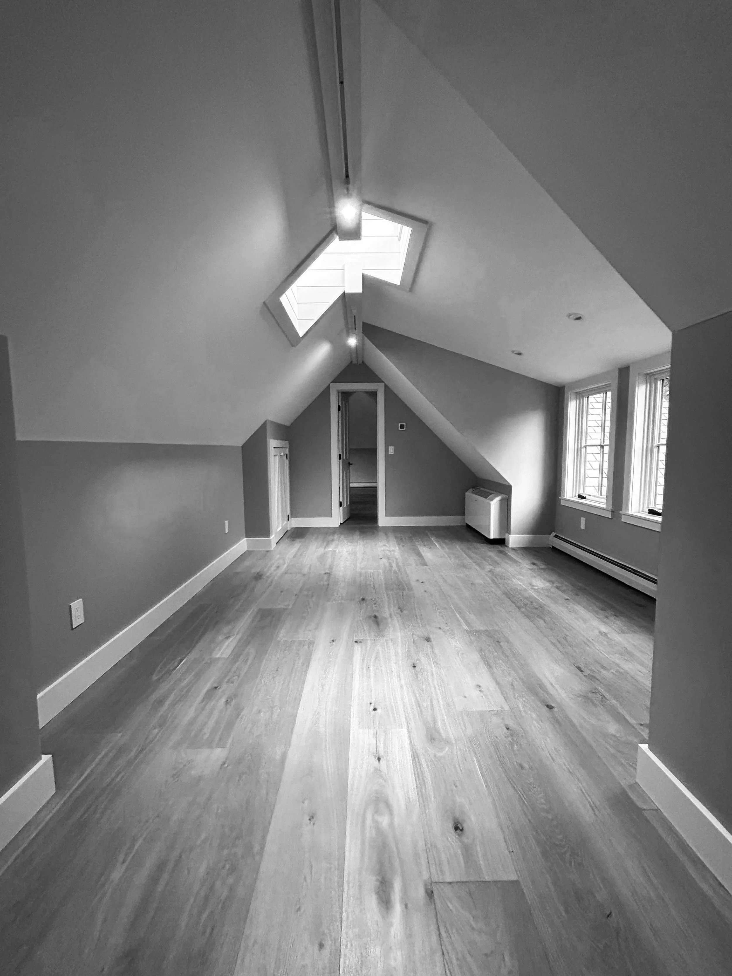 Empty attic room with sloped ceilings, hardwood flooring, three windows, and a skylight, in black and white. Built by the Timber, Stone, & Steel lead contractor, Taylor Prouty, in Vermont.