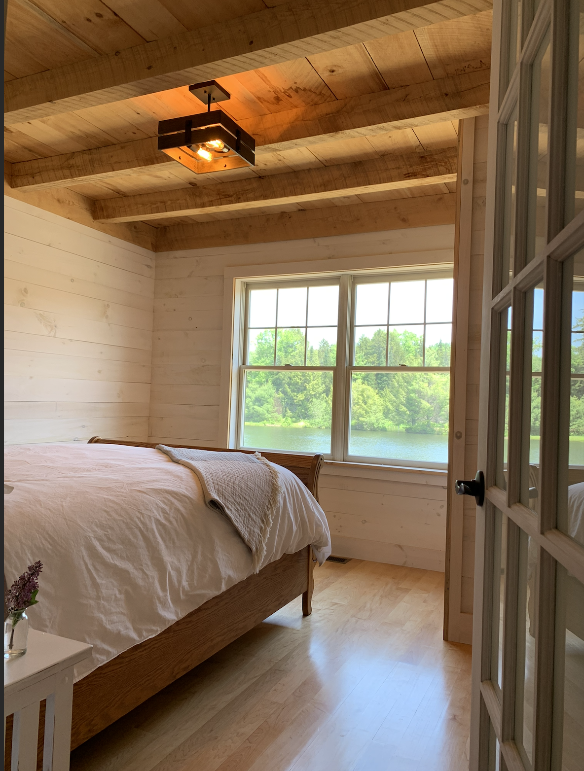 Bedroom with wooden walls and ceiling, large window showing a lake outside, bed with white bedding and beige blanket, small white nightstand with a vase of flowers, glass-paneled door. Built by Timber, Stone, & Steel, lead contractor in Vermont.