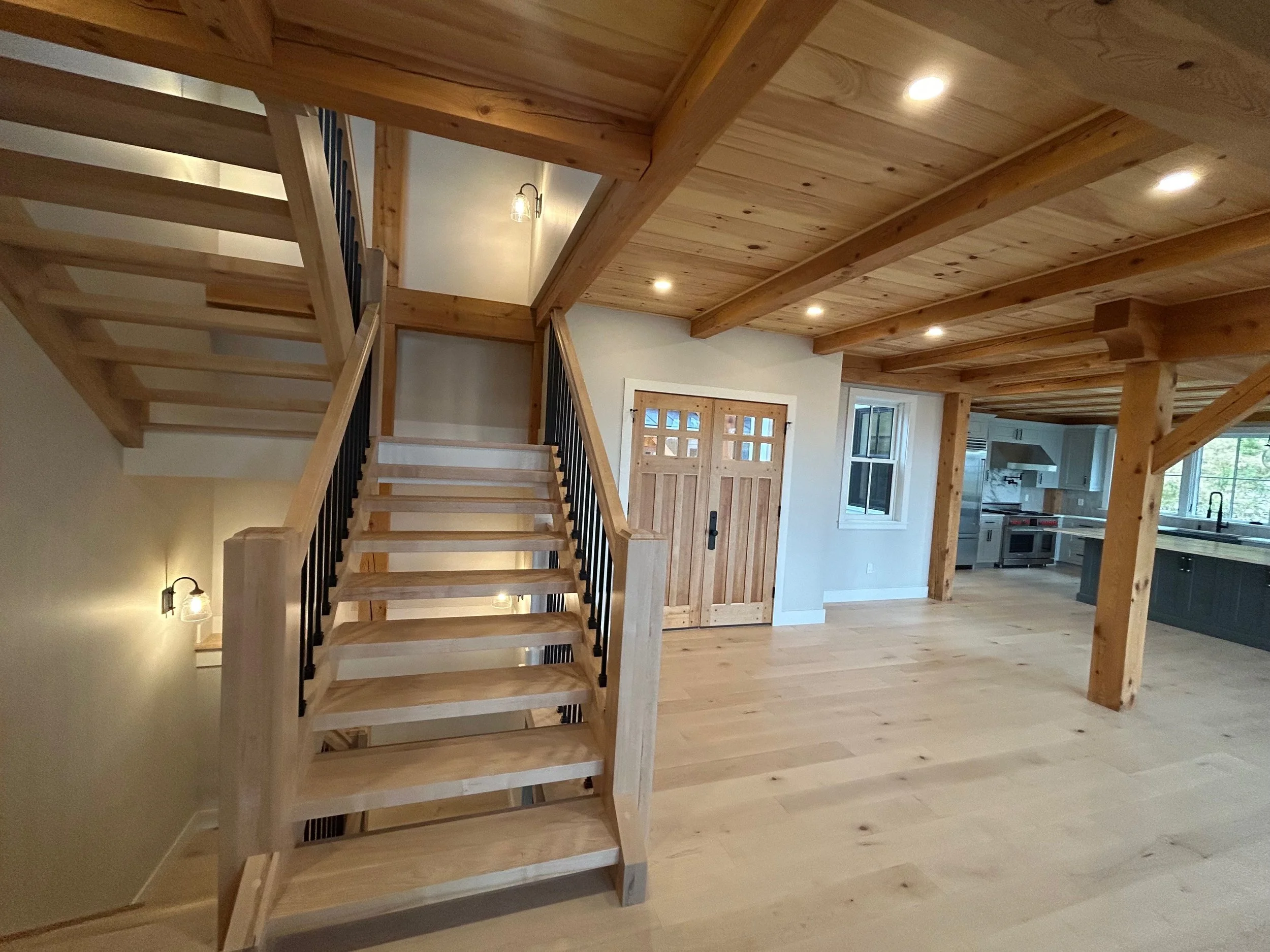 Open-concept modern home interior featuring a wooden staircase with black railings, wooden beams, and a kitchen area with stainless steel appliances and large windows. Built by the Timber, Stone, & Steel lead contractor, Taylor Prouty, in Vermont.