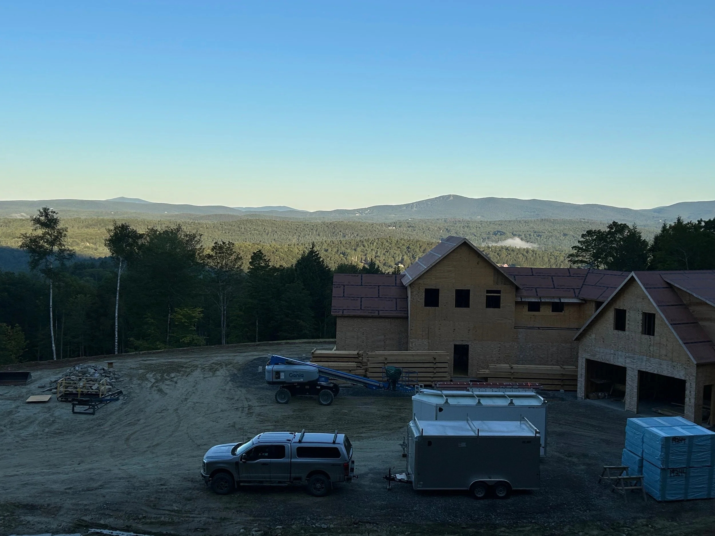 Construction site with partially built houses, construction vehicles, and supplies, set against a backdrop of trees and mountains under a clear blue sky. Built by the Timber, Stone, & Steel lead contractor, Taylor Prouty, in Vermont.