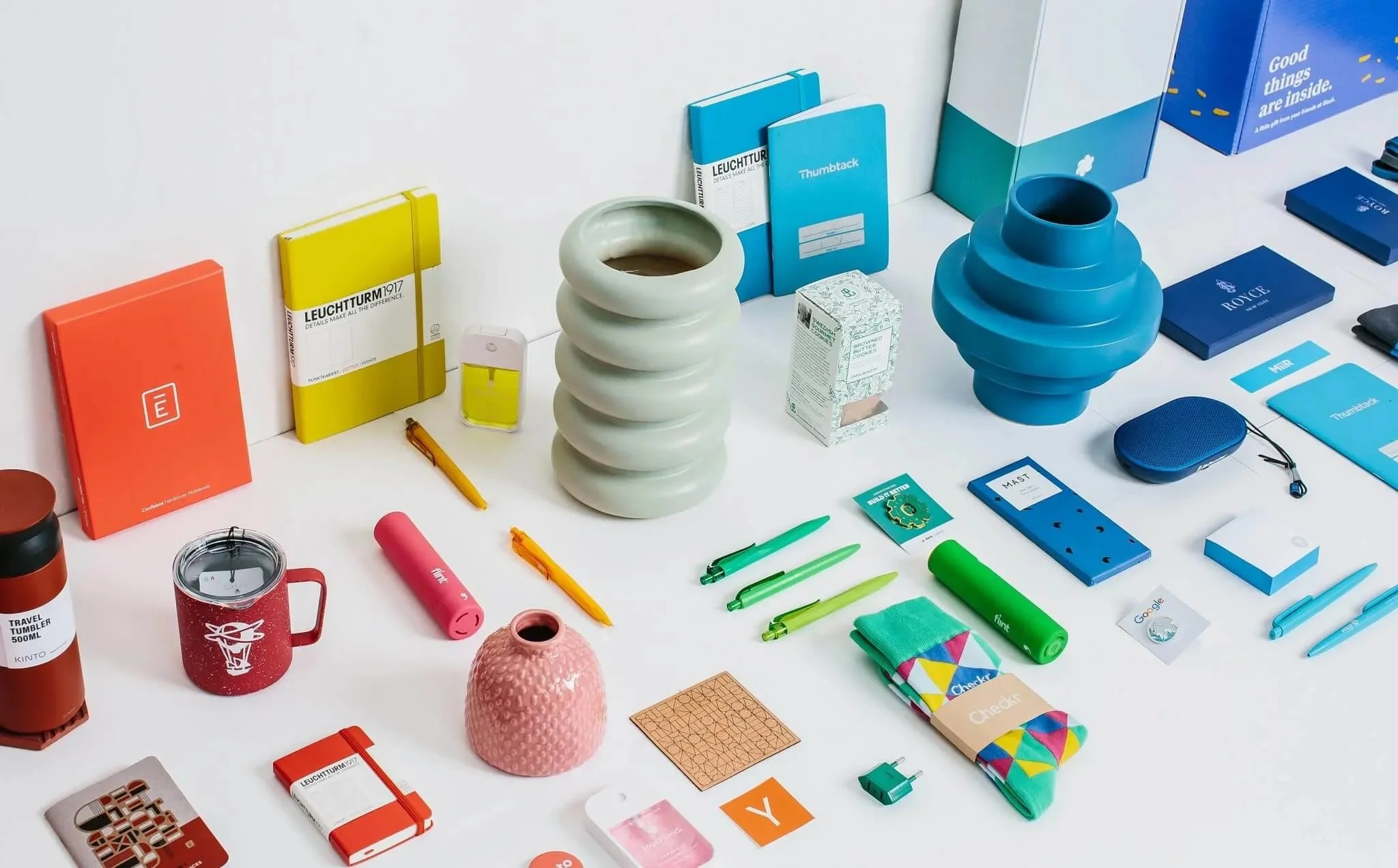 A variety of colorful office and travel accessories, including notebooks, pens, a mug, a pink vase, speakers, small boxes, and decorative items, arranged on a white surface to show the various options of promotional products.
