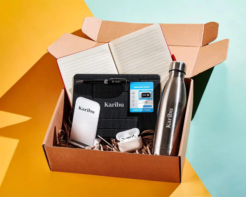 Open cardboard box containing a notebook, a black notebook cover with a pen, a wireless charger, a stainless steel  water bottle, a pair of wireless earbuds, and a small boxed webcam cover, showing an example kit for fullfillment.