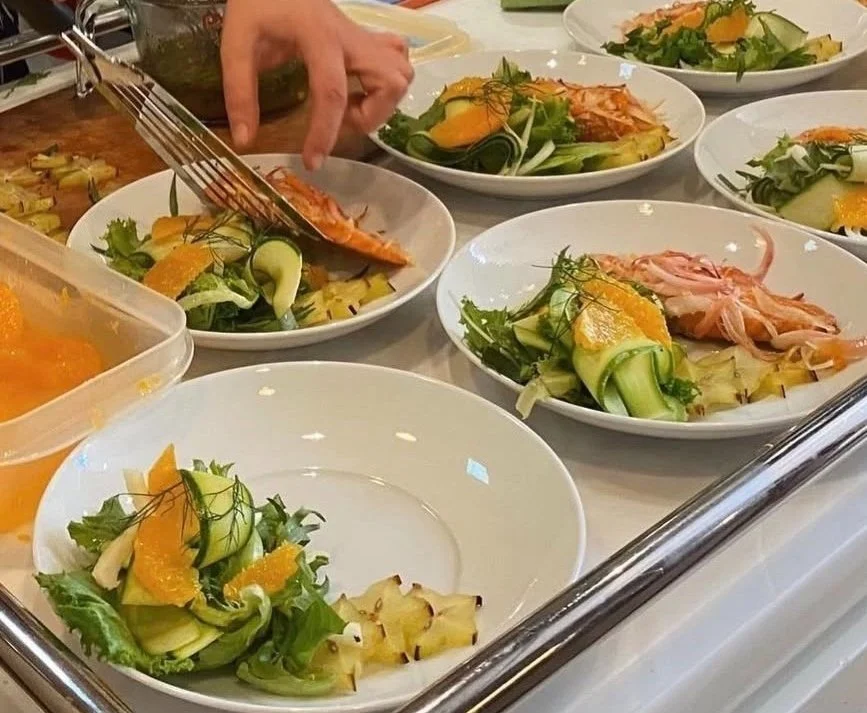 Plating slow cooked citrus salmon with red onion, featuring a salad of sliced fennel, courgette ribbons, orange segments, star fruit and fennel fronds. A hand is adding grilled salmon to one of the plates.