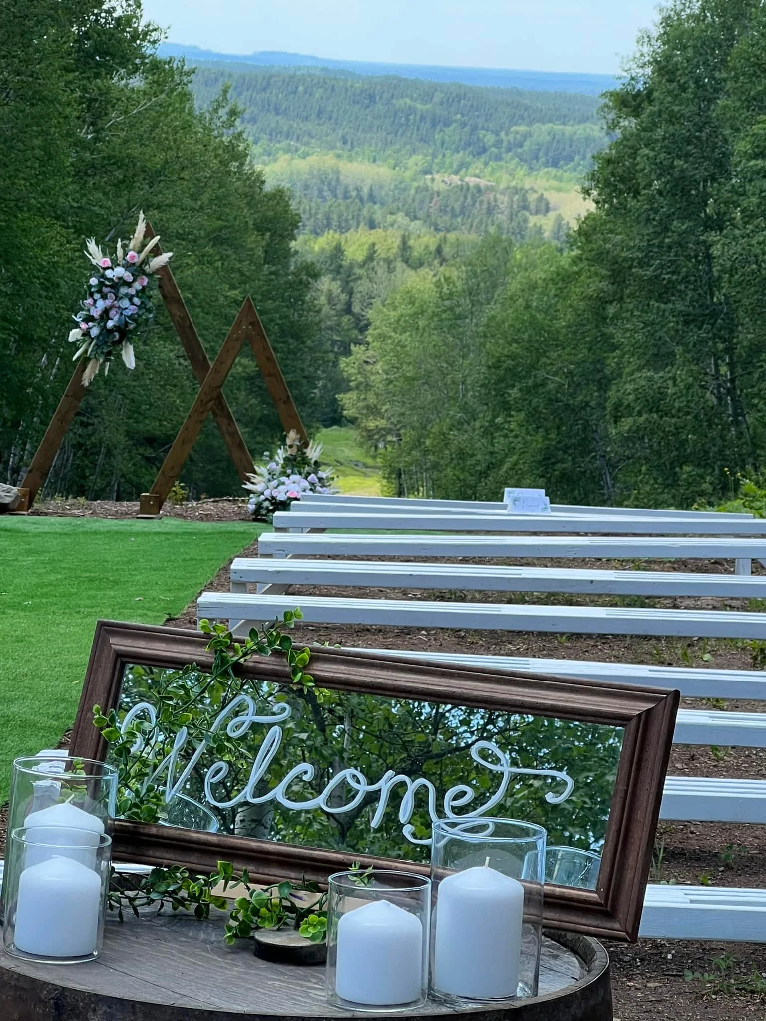 Outdoor wedding setup in a forested area with white benches, a wooden arbor decorated with flowers, and a welcome sign with candles in glass holders.