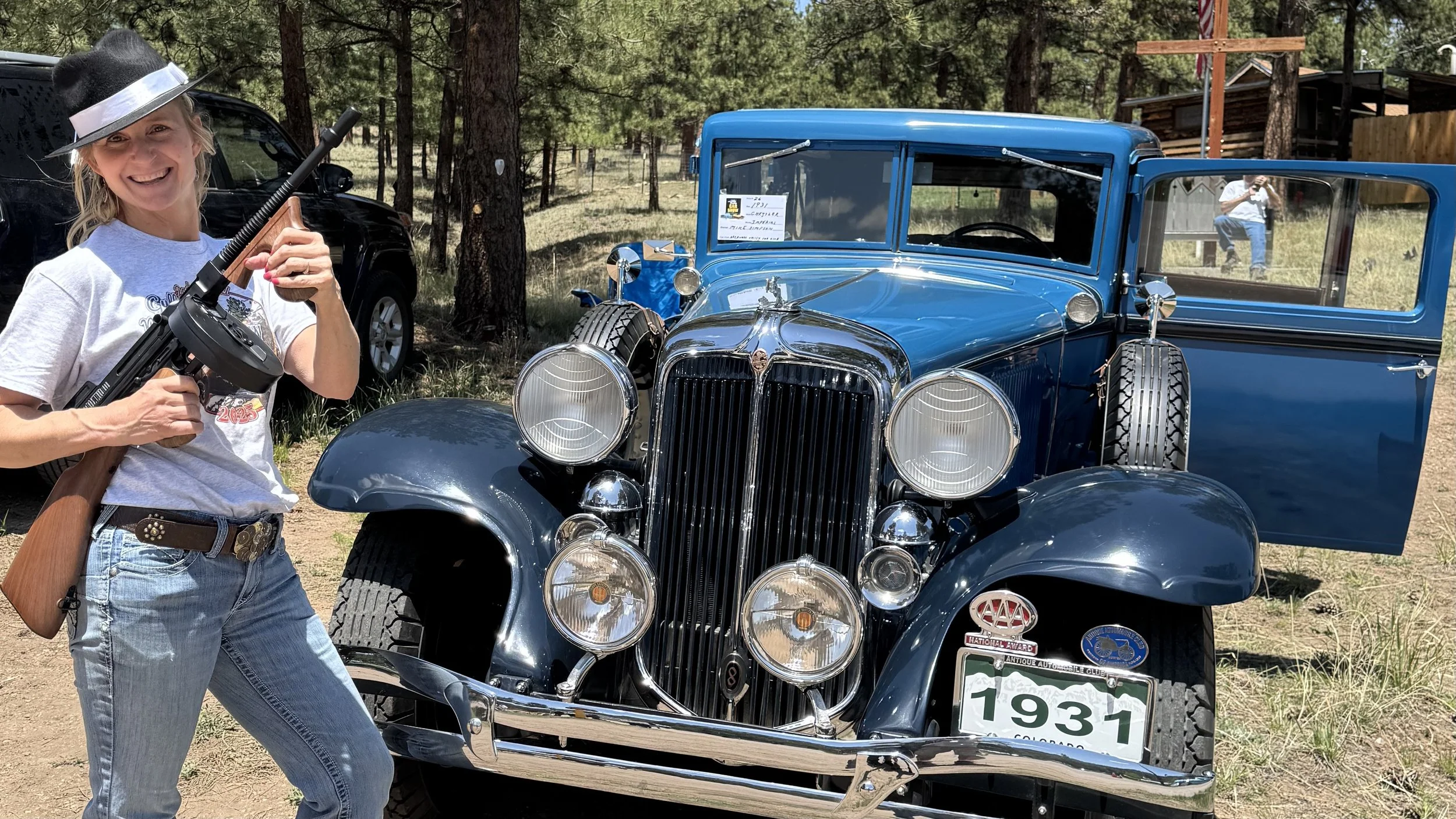 A woman smiling and holding a toy machine gun in front of a vintage blue car with a Colorado license plate from 1931, parked outdoors among trees.