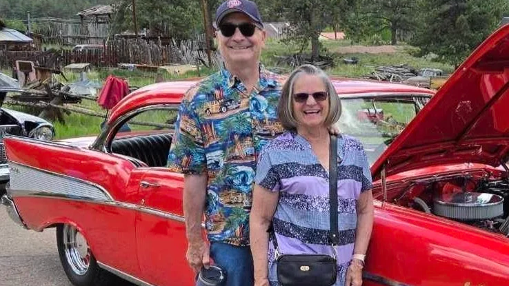 A smiling older man and woman standing next to a red vintage car with an open hood, outdoors in a rural area.