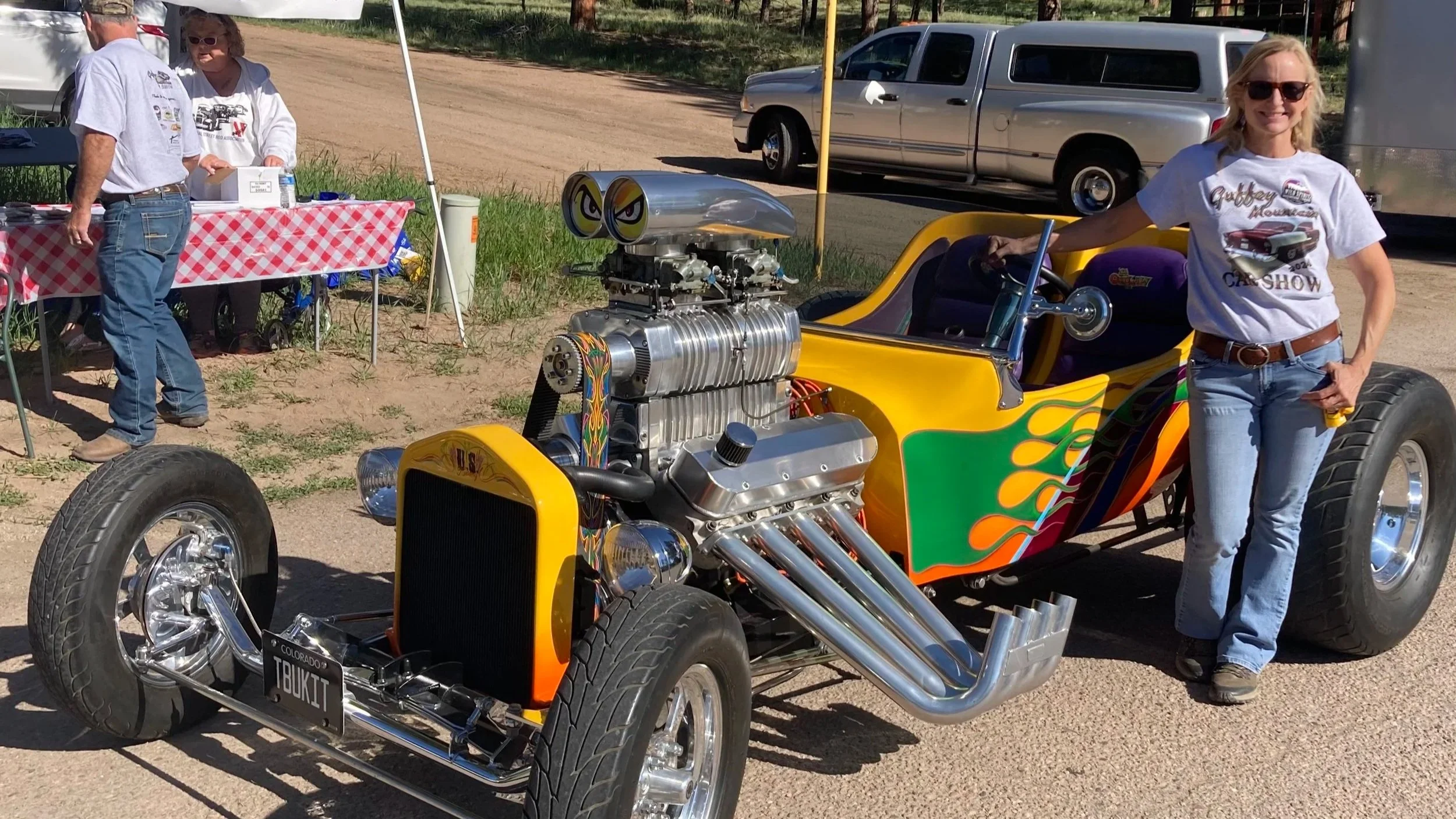 A woman standing next to a colorful vintage hot rod car with a large engine and flames painted on the side at an outdoor event. There is a registration table with two people in the background and a parked truck nearby.